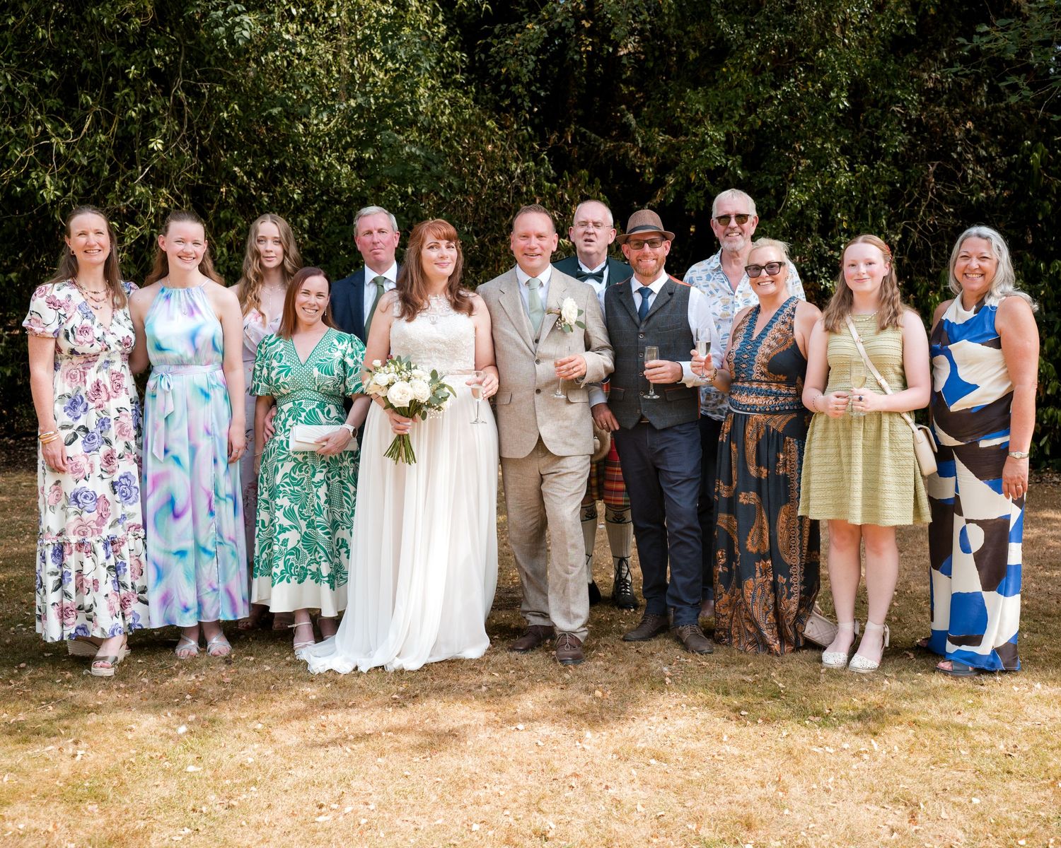 Wedding party group photo outdoors with bride in white gown, groom, and guests in formal attire, posing before lush green trees.