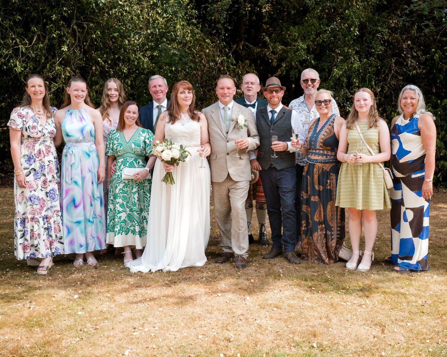 Wedding group photo with bride in white gown and groom in tan suit surrounded by smiling family outdoors.