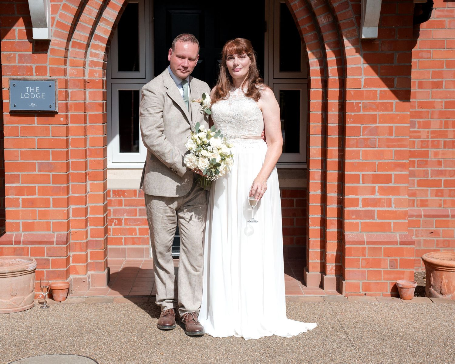 Bride and groom posing together outside a red brick building on their wedding day, bride holding white floral bouquet.