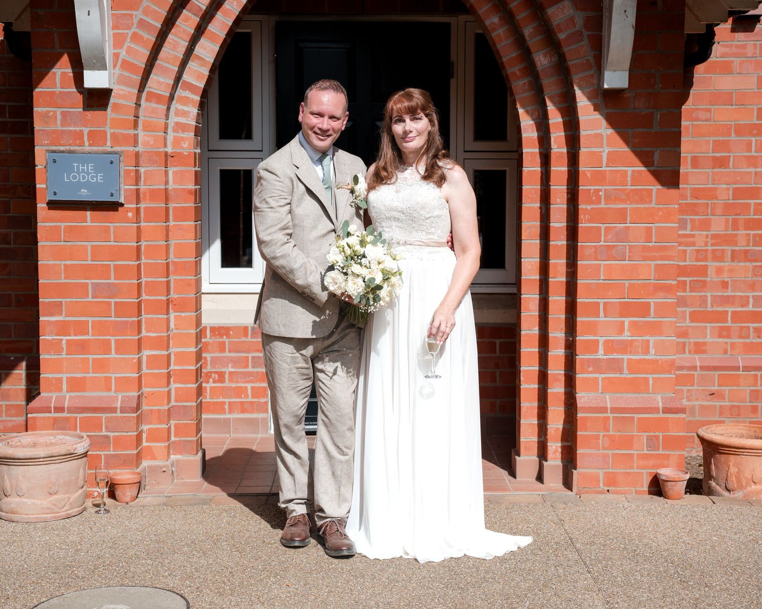 Bride and groom posing together outside a red brick church, bride holding white bouquet, groom in tan suit.