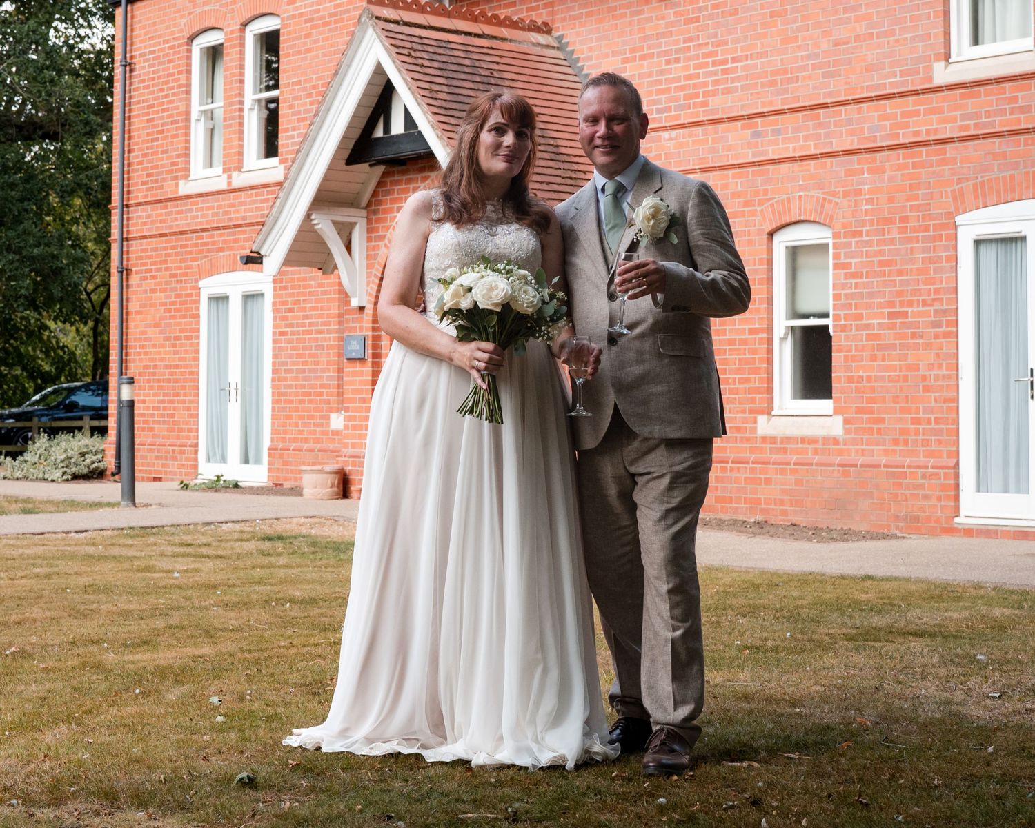 Bride in white gown and groom in tan suit pose together outside a red brick building on their wedding day.