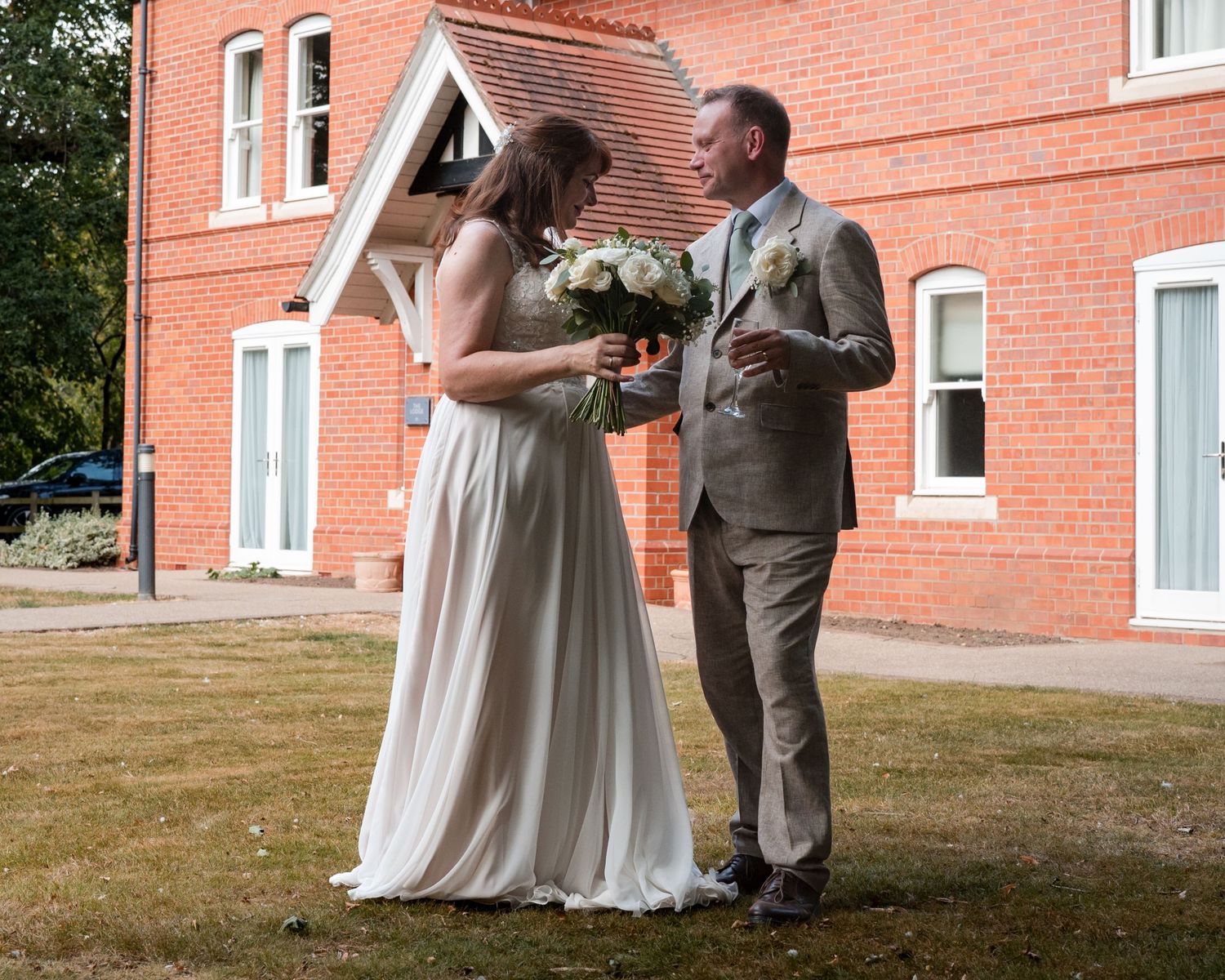 Bride in white gown holds bouquet with groom in tan suit outside red brick building on wedding day.