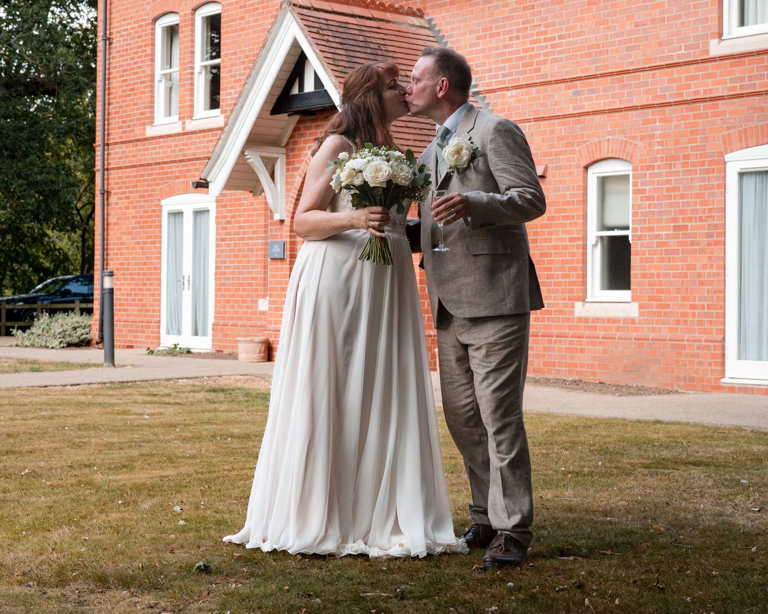 Bride and groom share an intimate moment outside a red brick venue, bride holding a white bouquet.