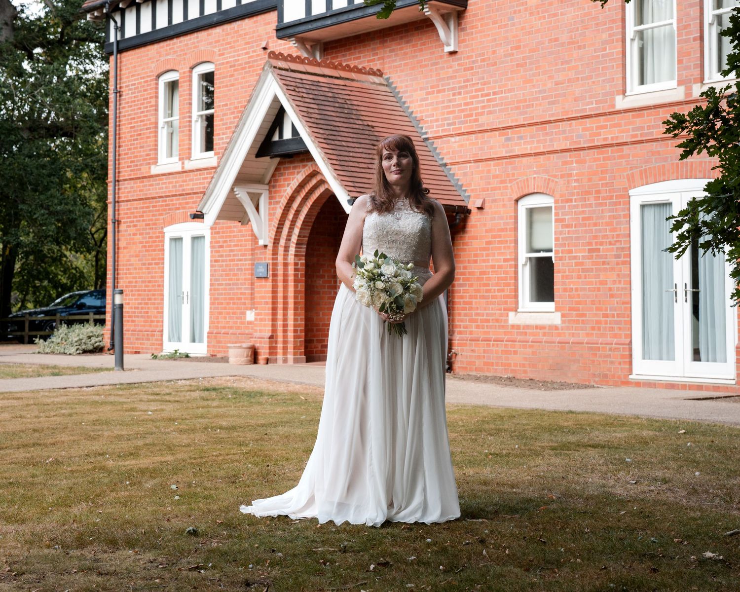 Bride in white gown holding bouquet stands before a red brick Victorian building on her wedding day.