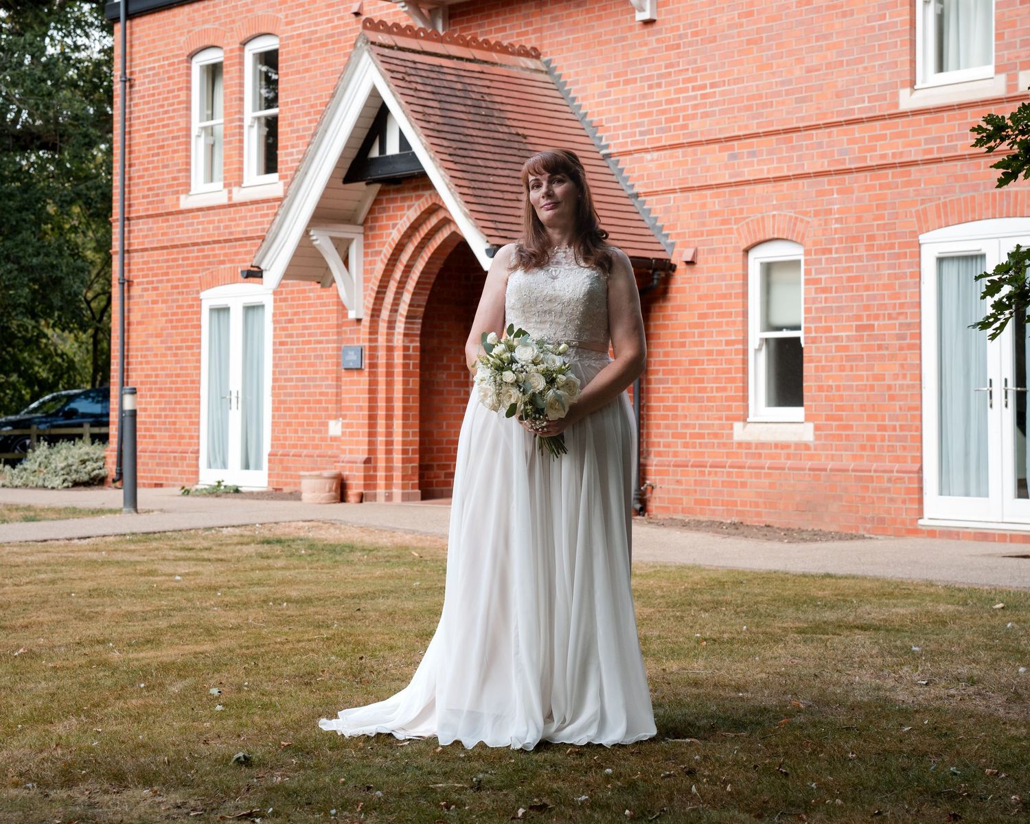 Bride in white wedding gown holding bouquet stands before red brick venue with arched entrance.