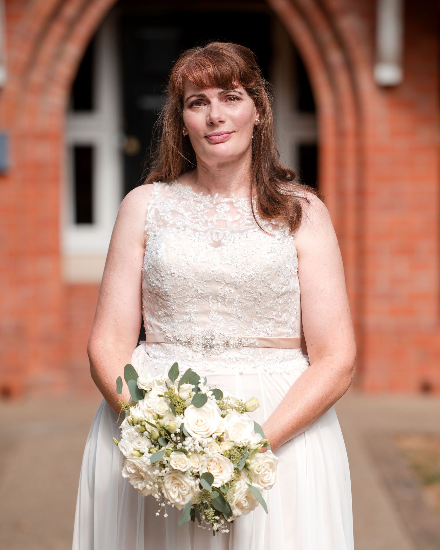 Bride in lace wedding dress holding white rose bouquet stands before brick building entrance.