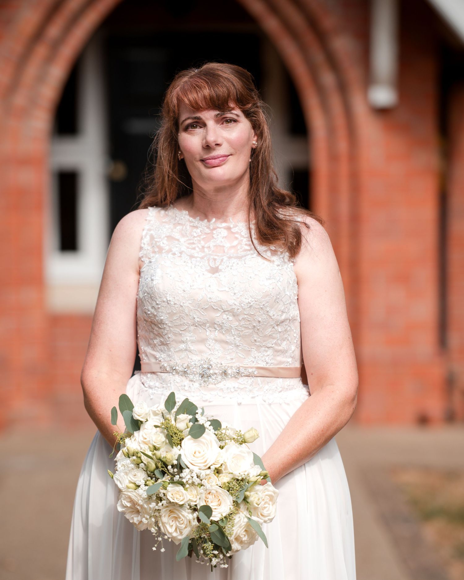Bride in lace wedding dress holding white rose bouquet stands before red brick church entrance.