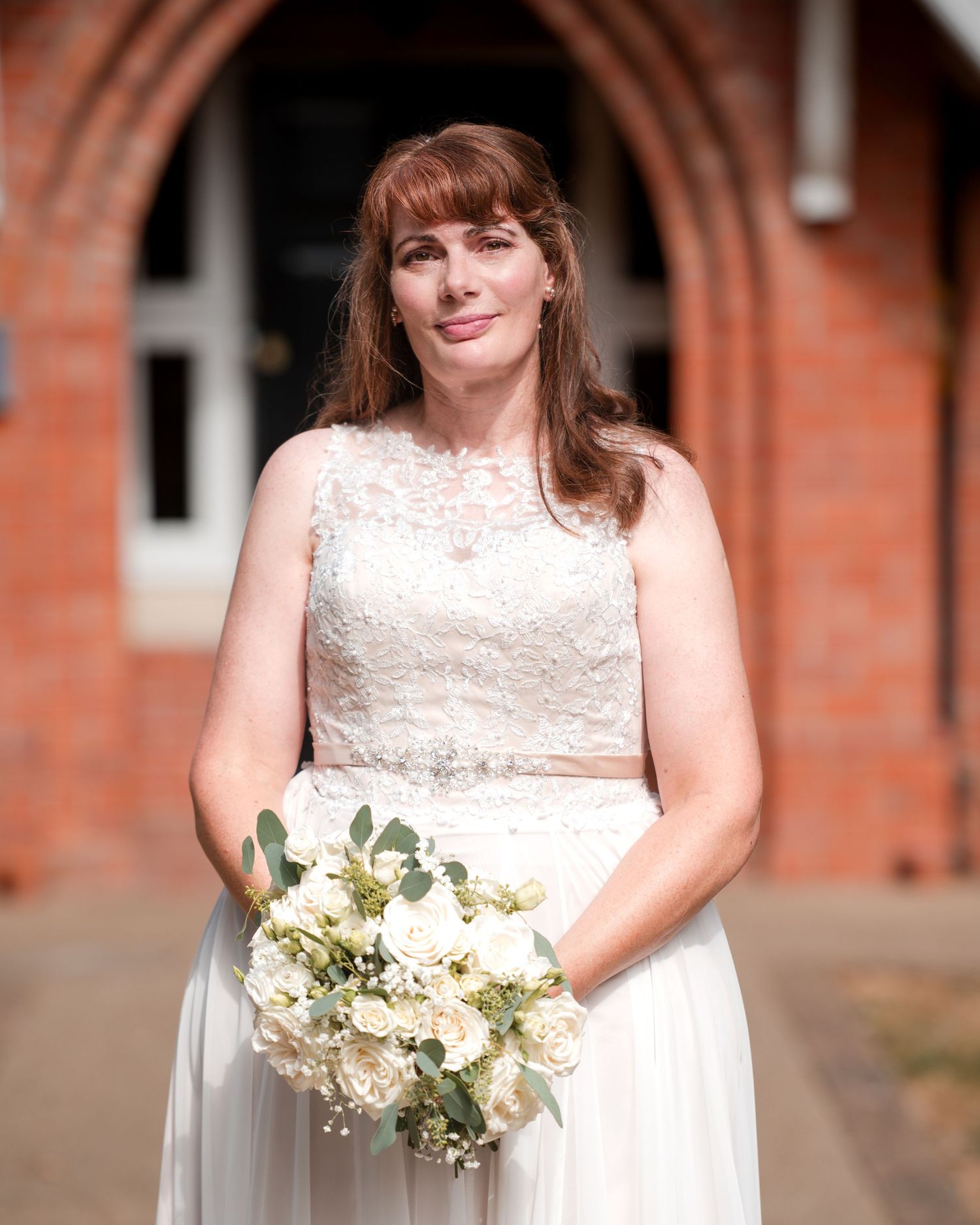 Bride in white lace wedding dress holding bouquet of white roses and eucalyptus outside brick church.