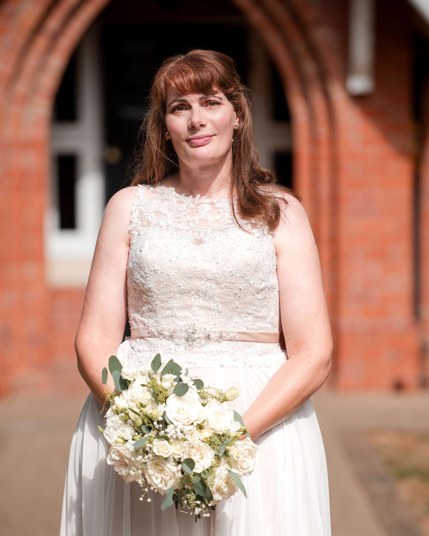 Bride in lace wedding dress holding white rose bouquet stands before brick church entrance.