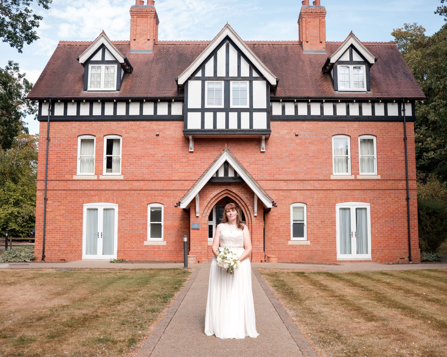 Bride in white gown holding bouquet stands before a grand Tudor-style red brick manor house on her wedding day.