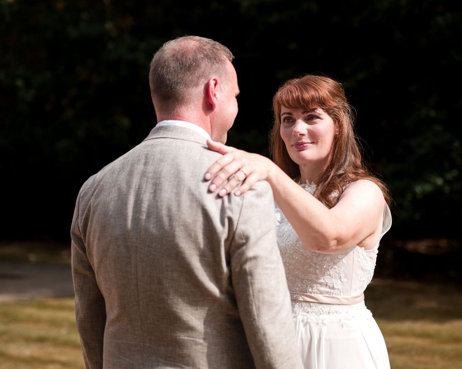 Bride in white dress places hand on groom's shoulder during outdoor wedding ceremony.