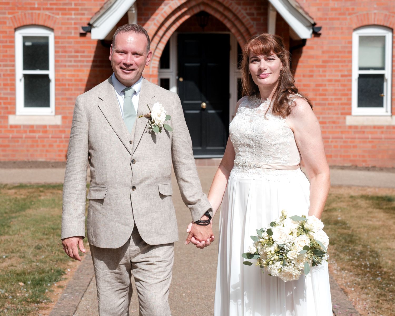 Bride and groom holding hands in front of a red brick church, bride holding white bouquet, groom wearing beige suit with boutonniere.