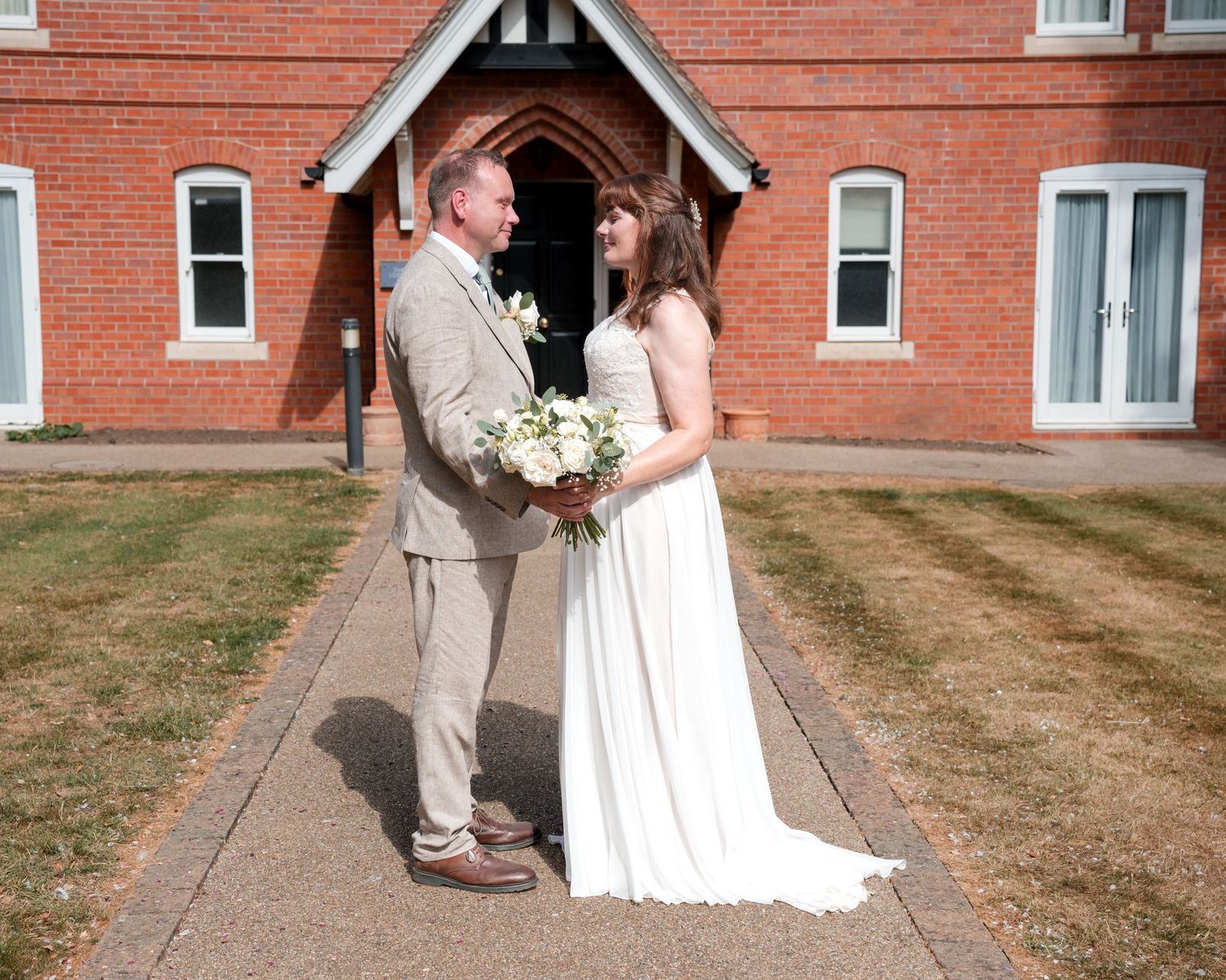 Bride and groom standing together outside a red brick church, bride holding white bouquet in elegant wedding dress.