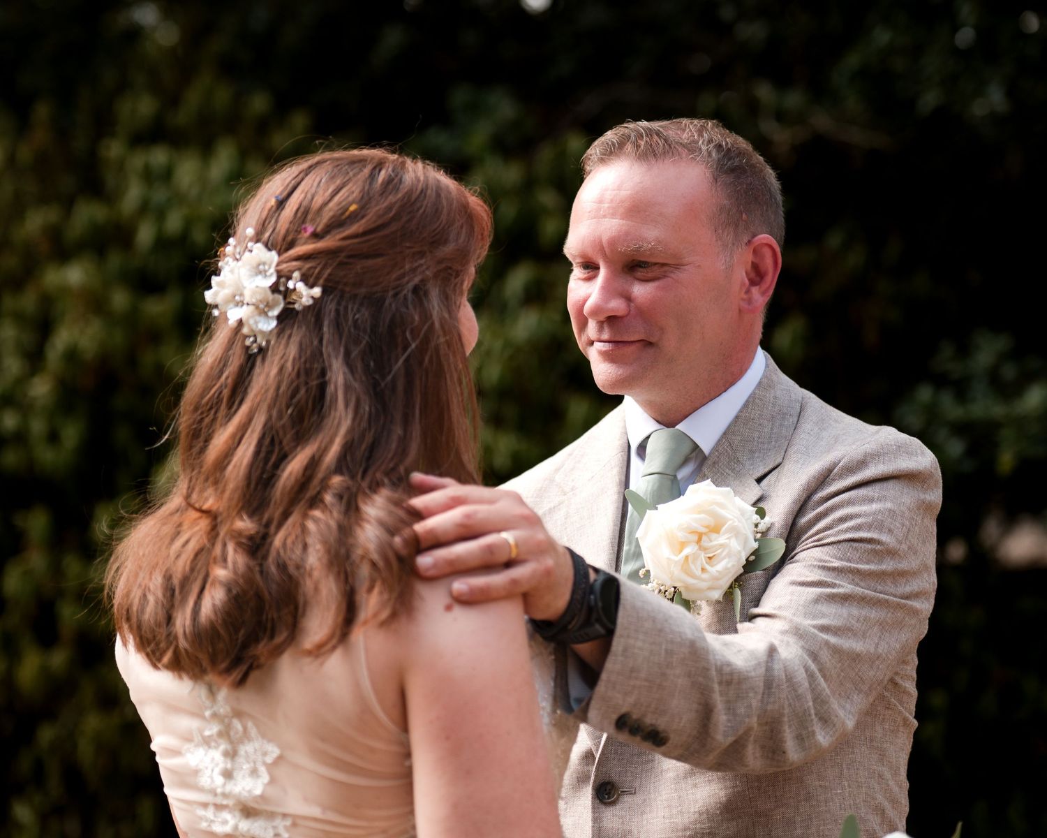 A groom in a tan suit smiles warmly at his bride outdoors, both wearing floral accessories at their wedding.