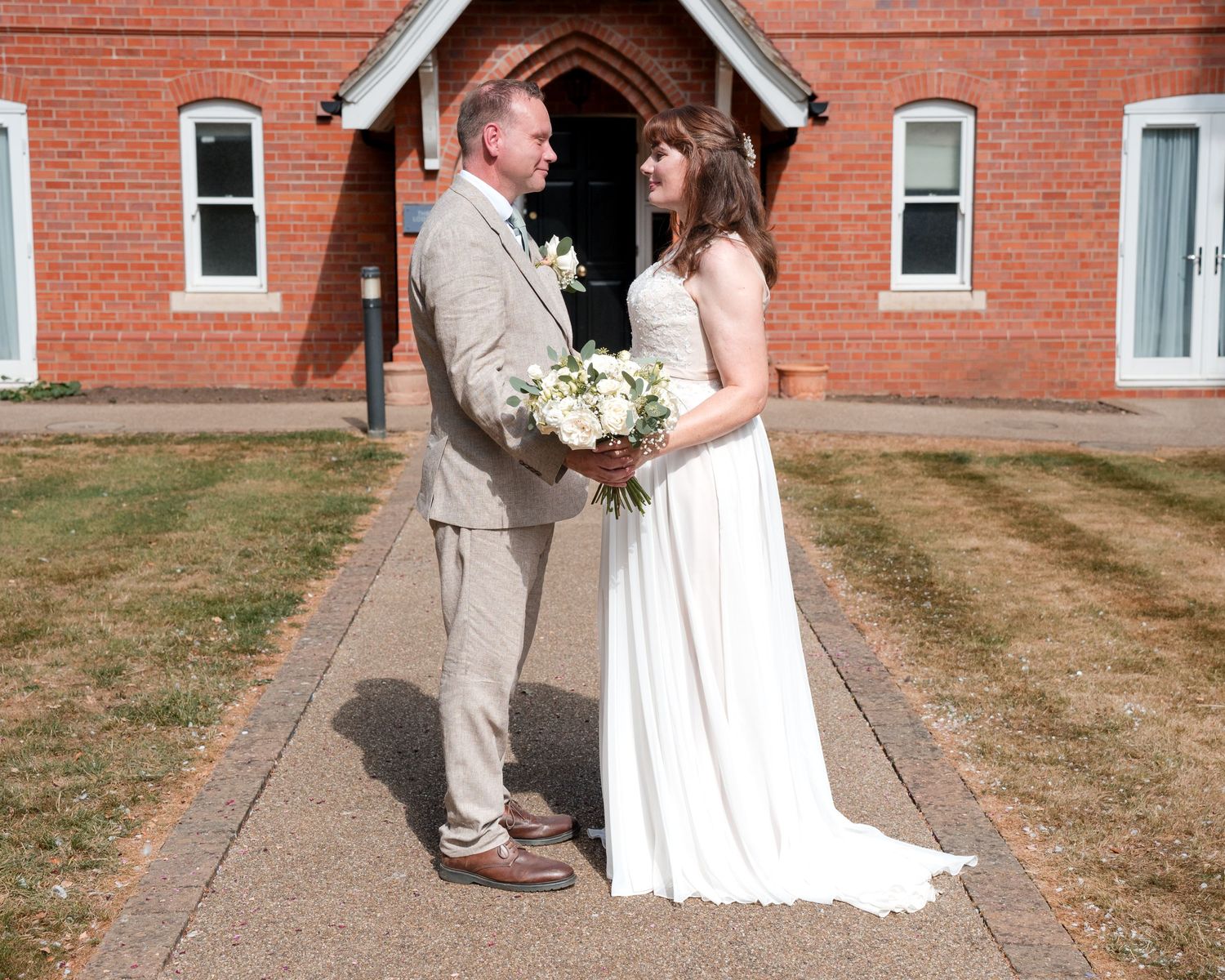 Bride and groom standing together on a church pathway, bride holding white bouquet, in front of a red brick chapel.