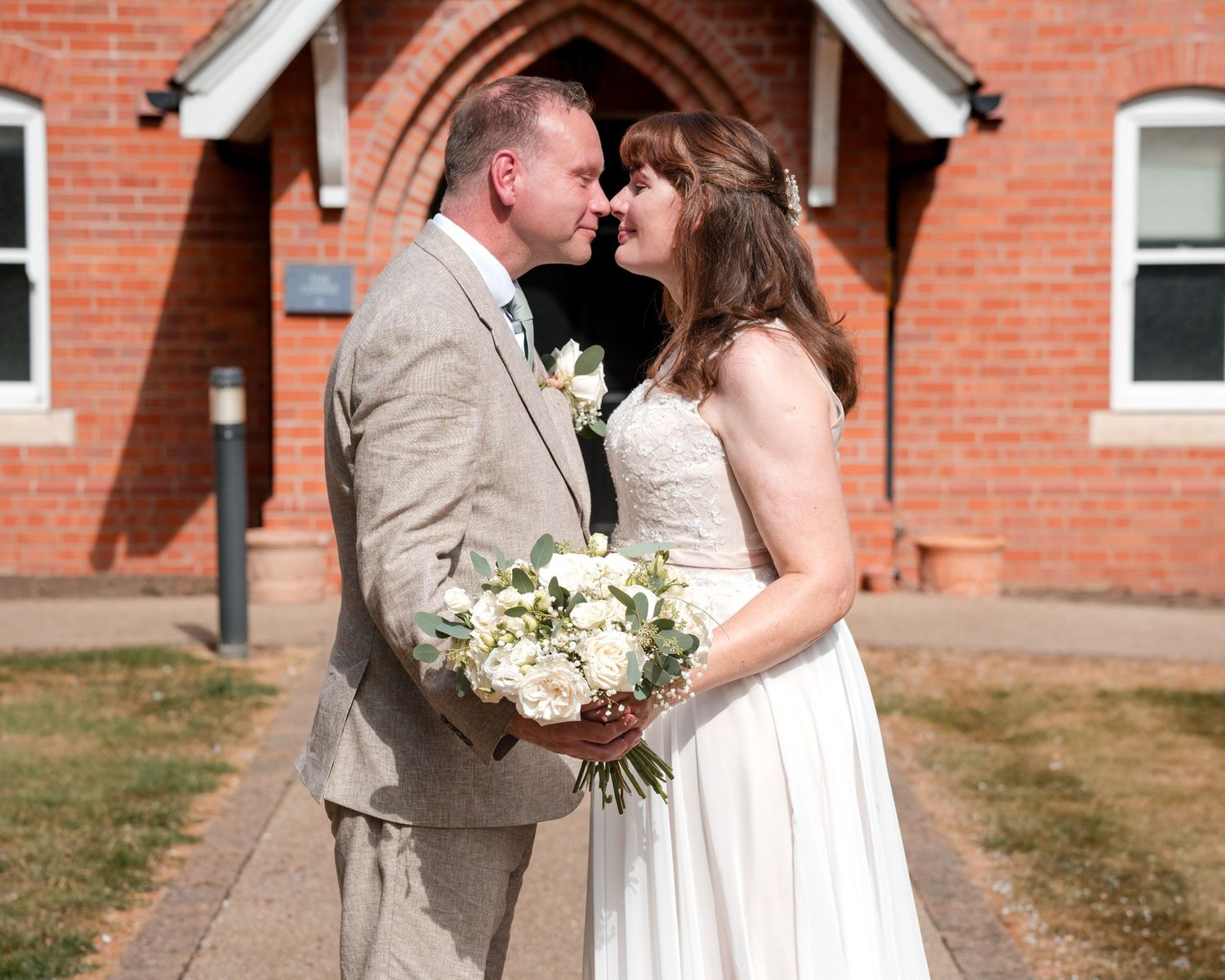 Bride and groom sharing a romantic moment outside a red brick church, bride holding white floral bouquet.