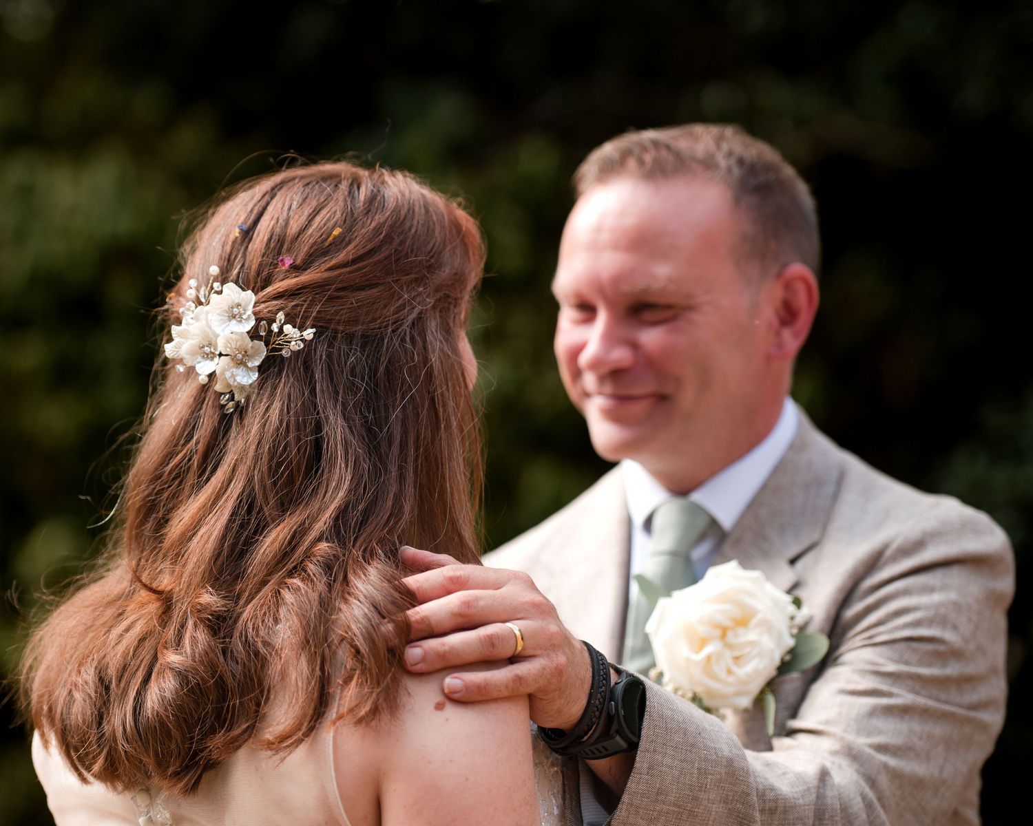 Groom in beige suit with white boutonniere smiling at bride with floral hair accessory at outdoor wedding.