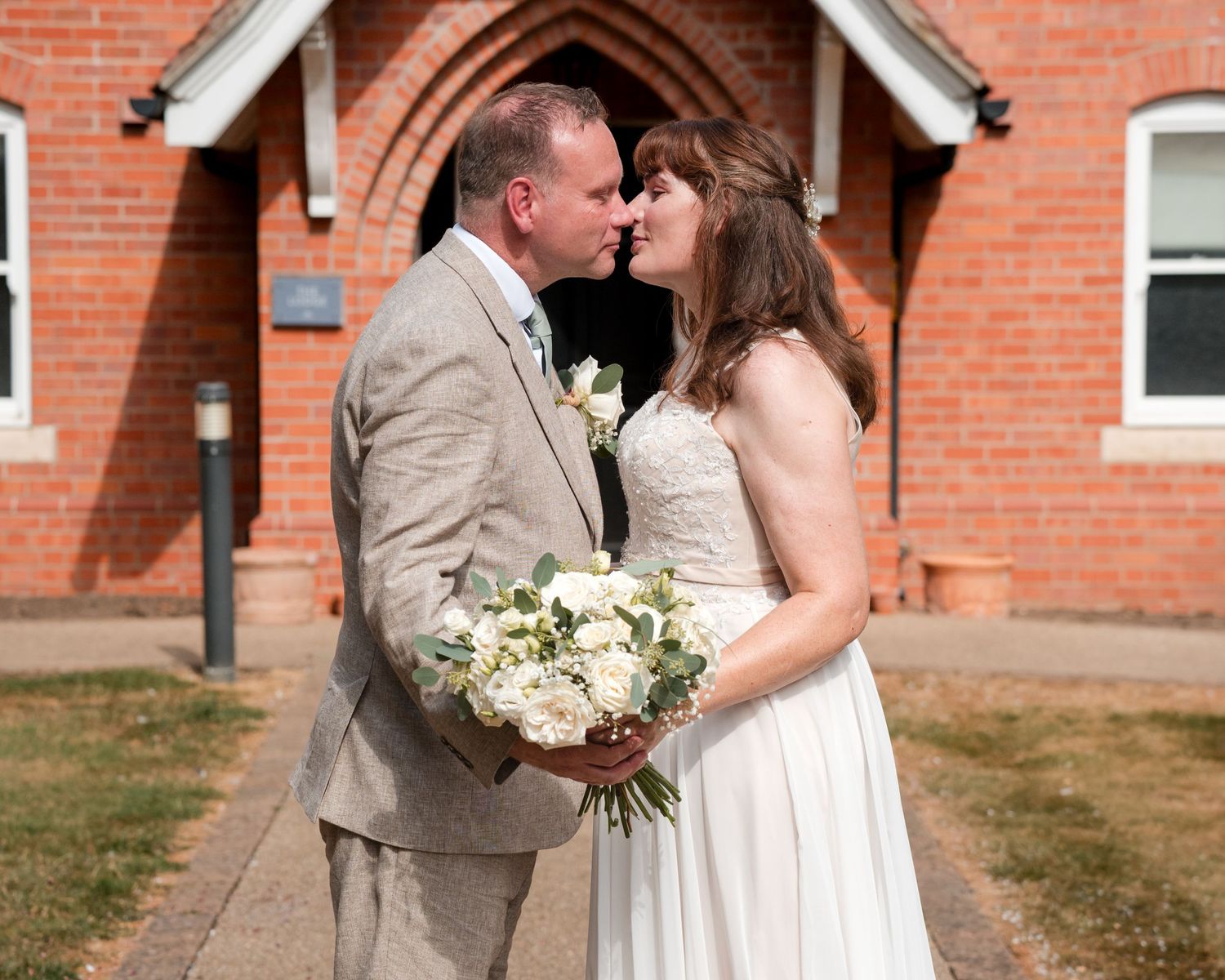 Bride and groom share a romantic kiss outside a red brick church, bride holding white floral bouquet.