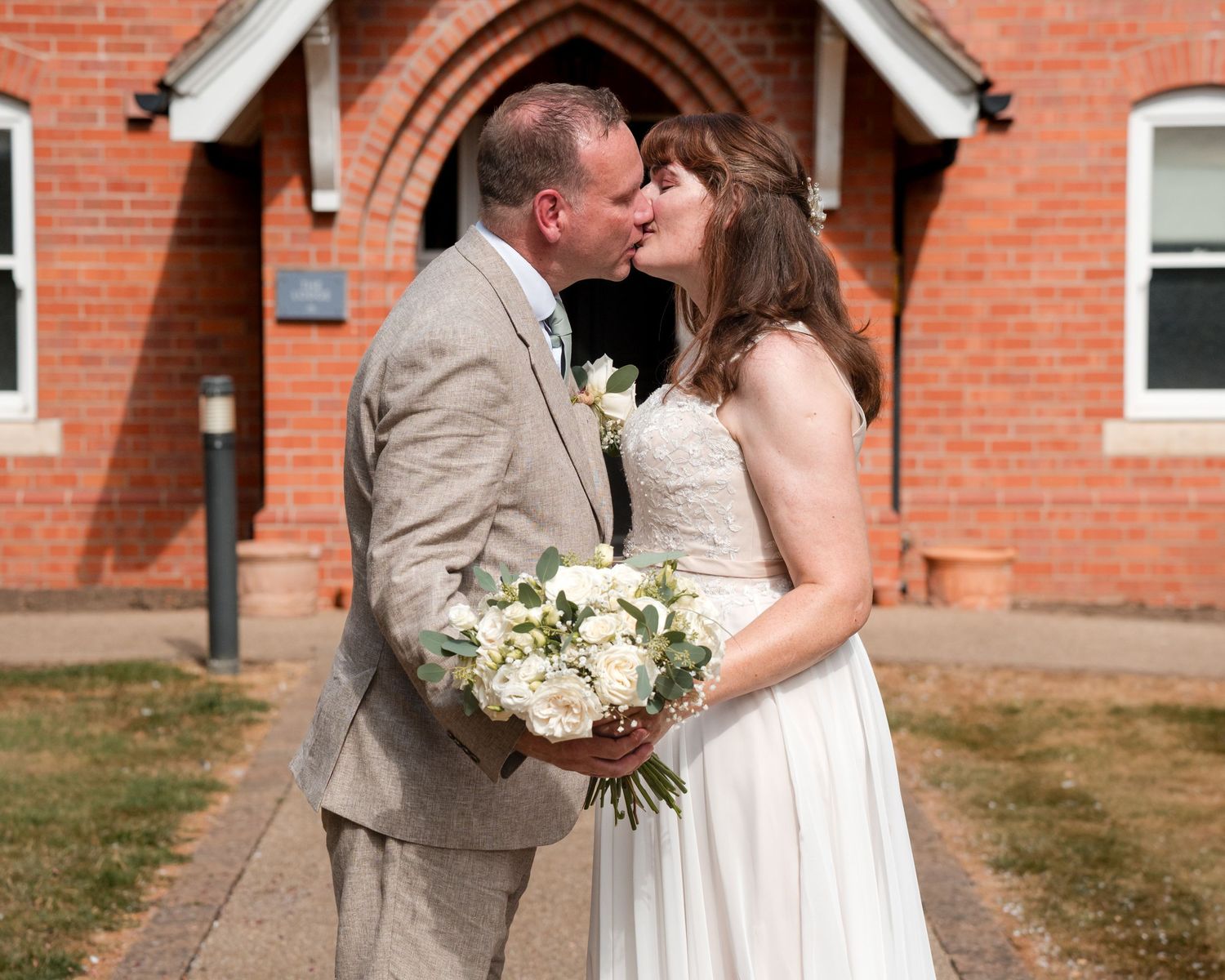 Bride and groom kissing in front of a red brick church, bride holding white floral bouquet in wedding attire.
