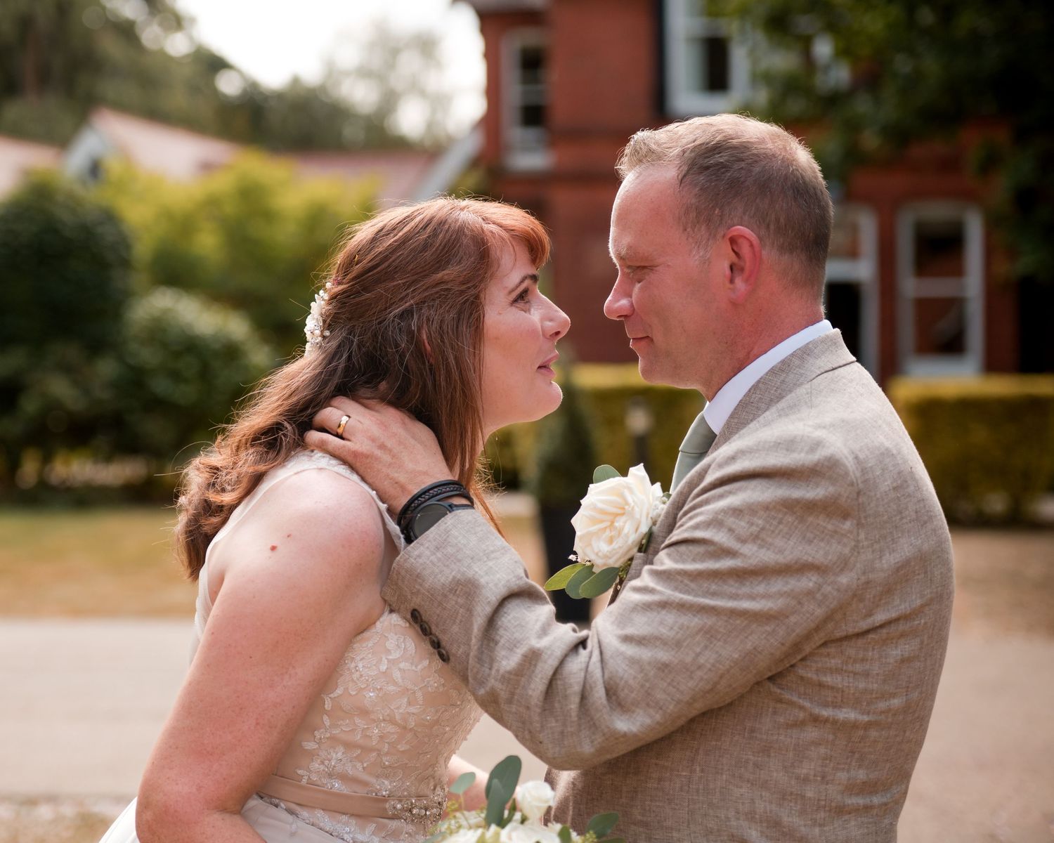 Bride and groom share a romantic moment outdoors, with the groom in a grey suit and bride in a lace wedding dress.