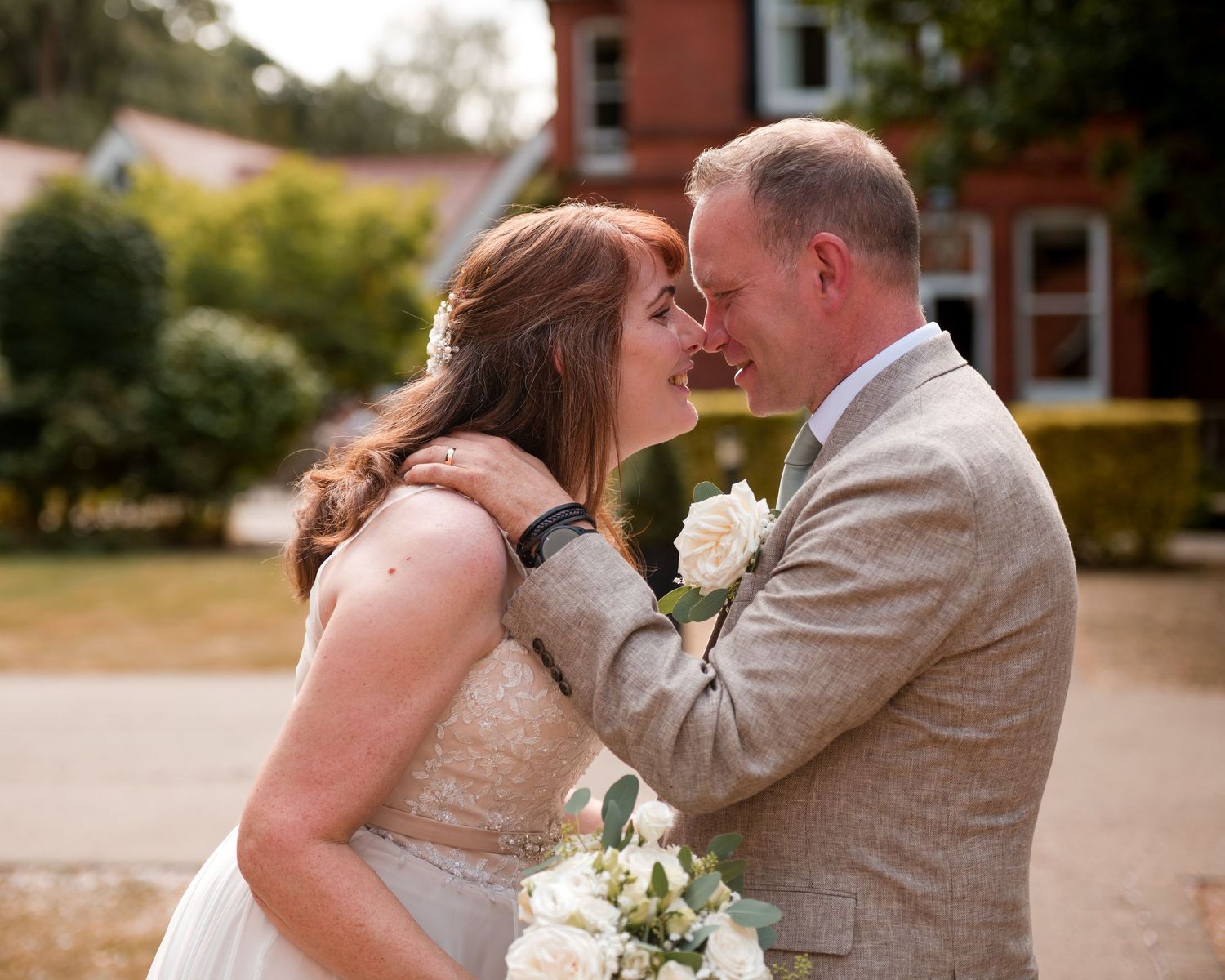 Bride and groom sharing a romantic moment, touching foreheads, bride holding white bouquet outside a red brick venue.