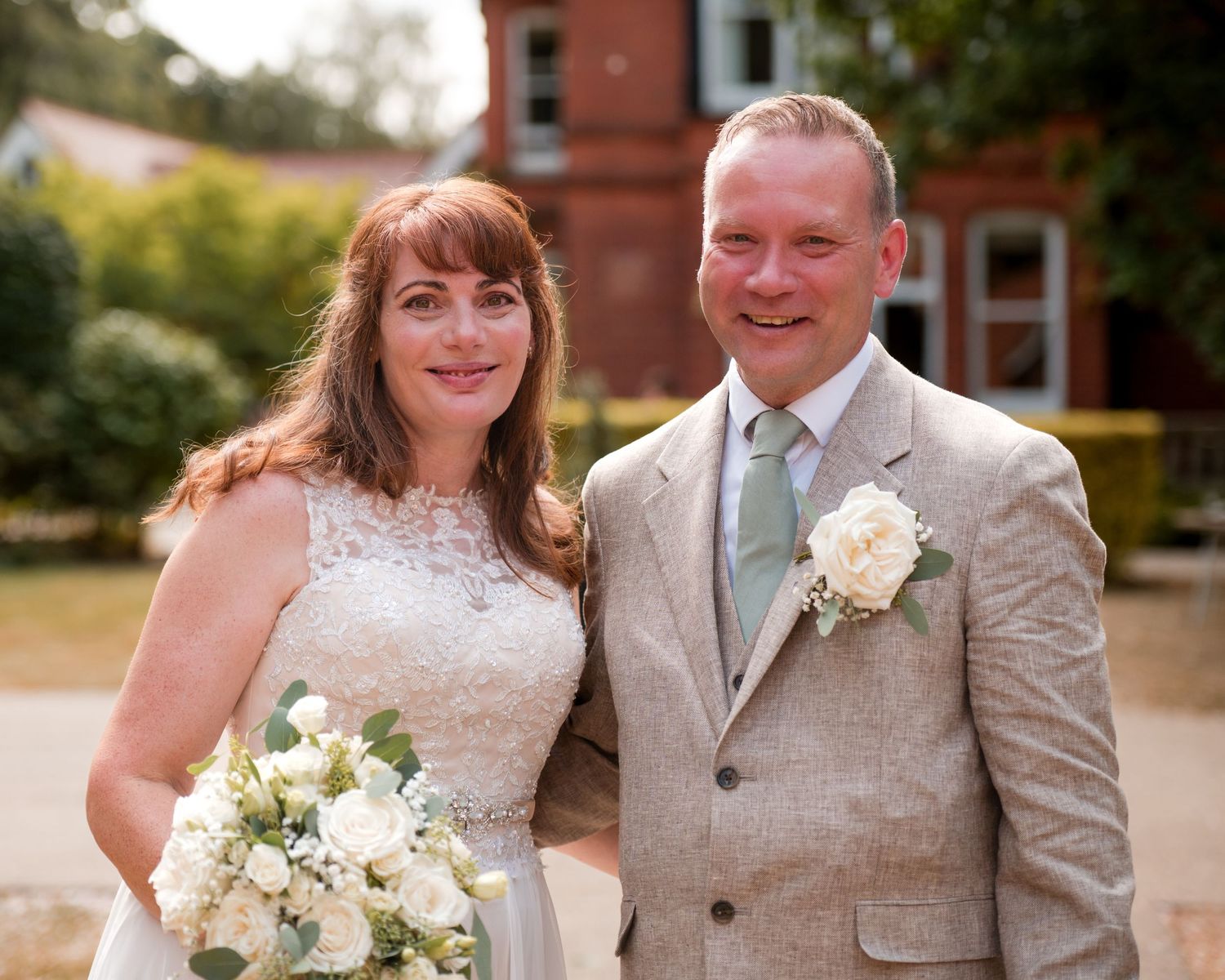 Smiling bride and groom posing outdoors on their wedding day, bride holding white bouquet, groom in tan suit with boutonniere.