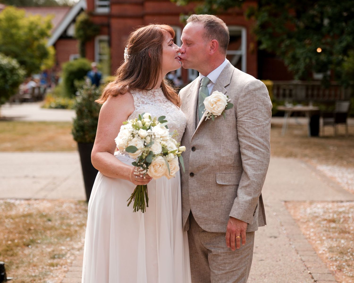 Bride and groom sharing a kiss outdoors, bride holding white bouquet, groom in beige suit with boutonniere.