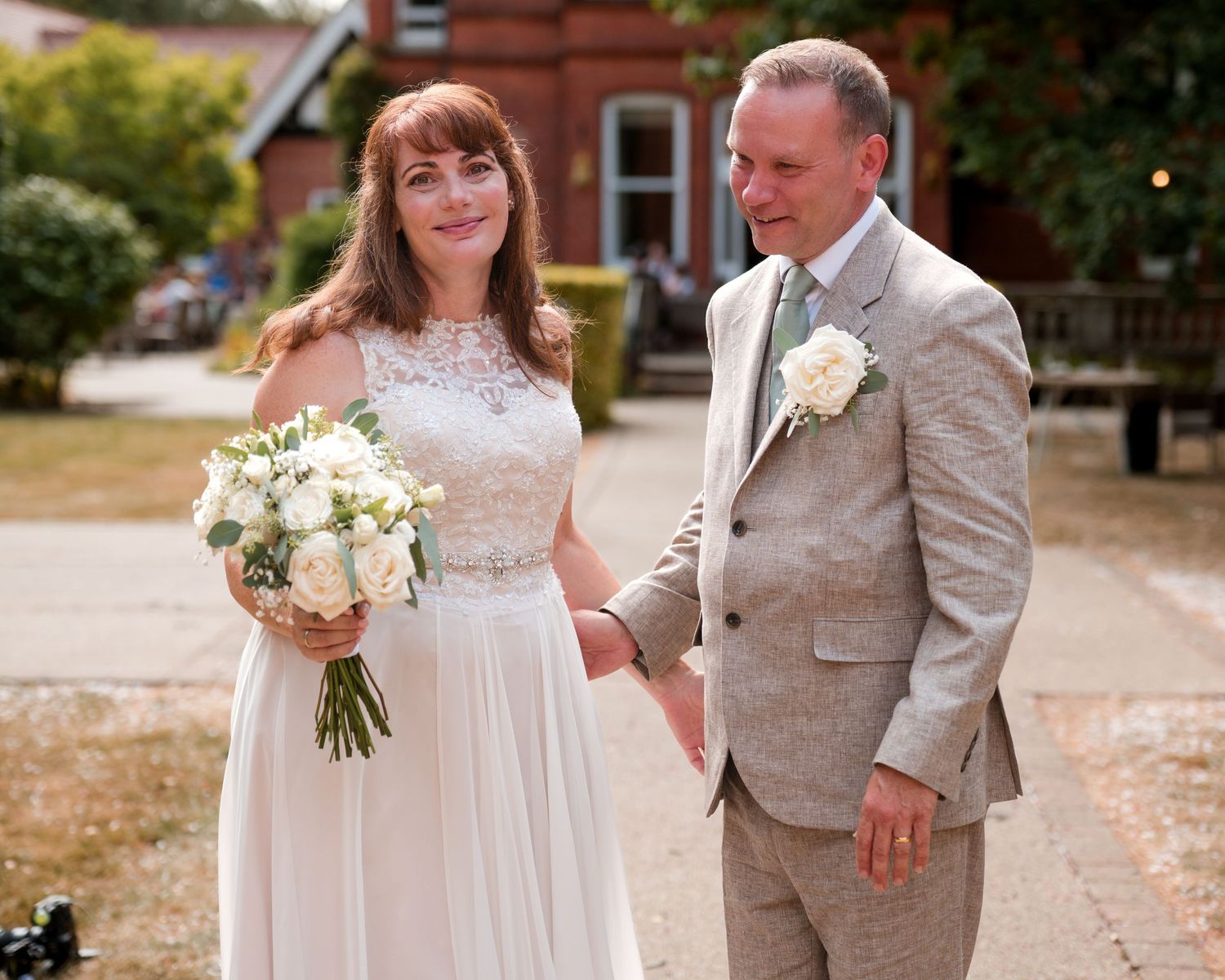 Bride in white gown holding bouquet stands with groom in tan suit at outdoor wedding venue.