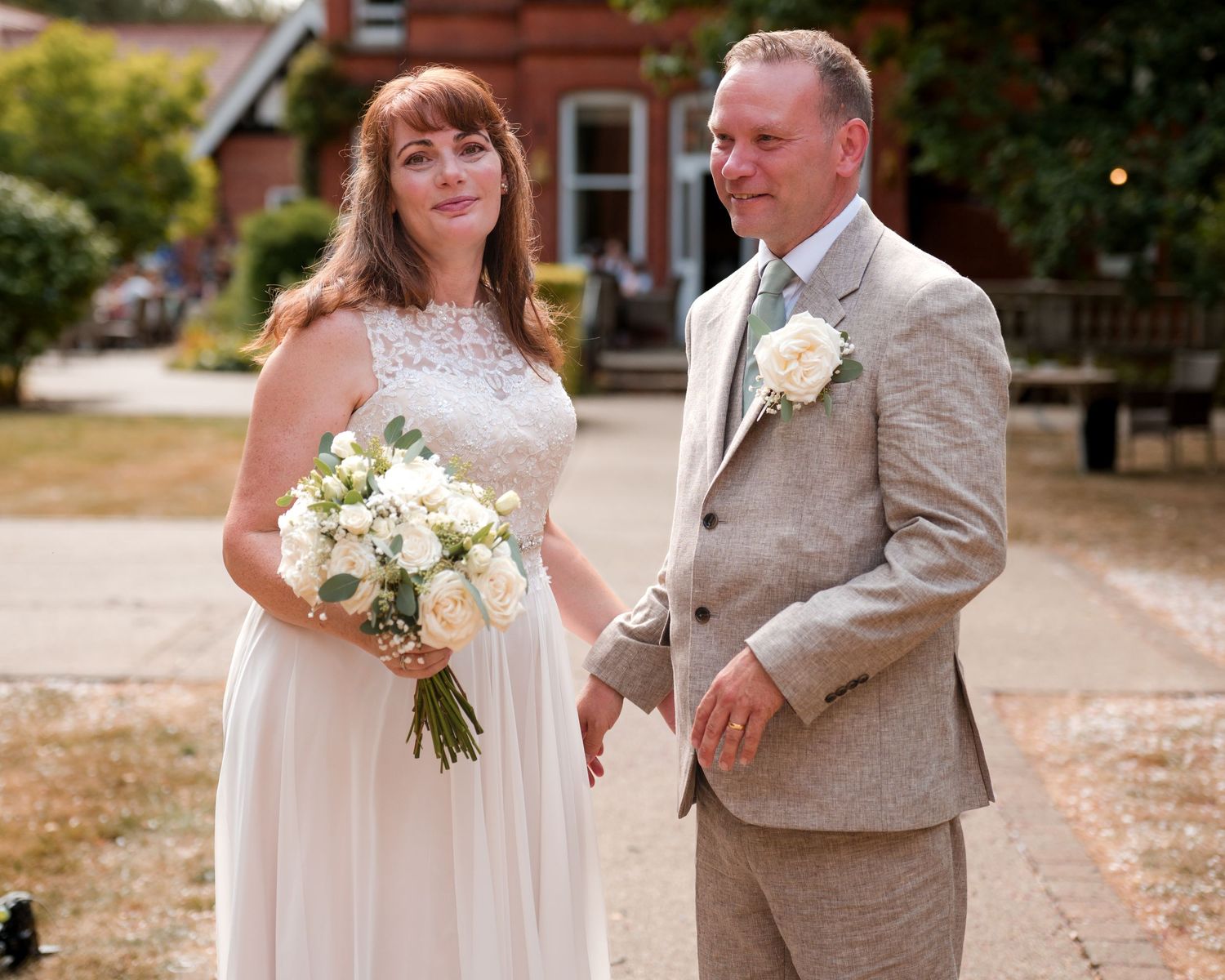 Bride and groom posing outdoors on their wedding day, bride holding white bouquet, groom in grey suit.