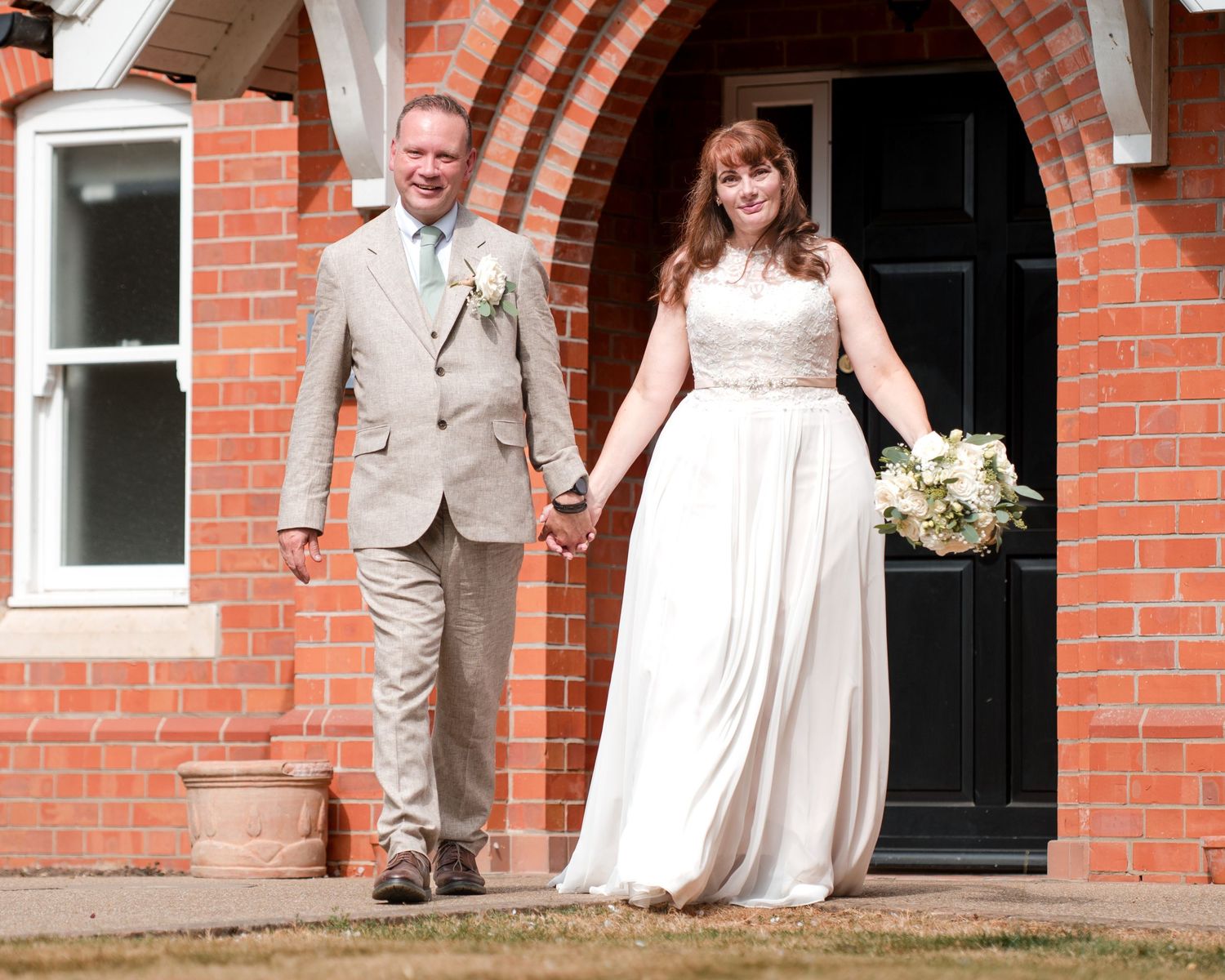 Bride in white gown and groom in tan suit hold hands outside a red brick church on their wedding day.