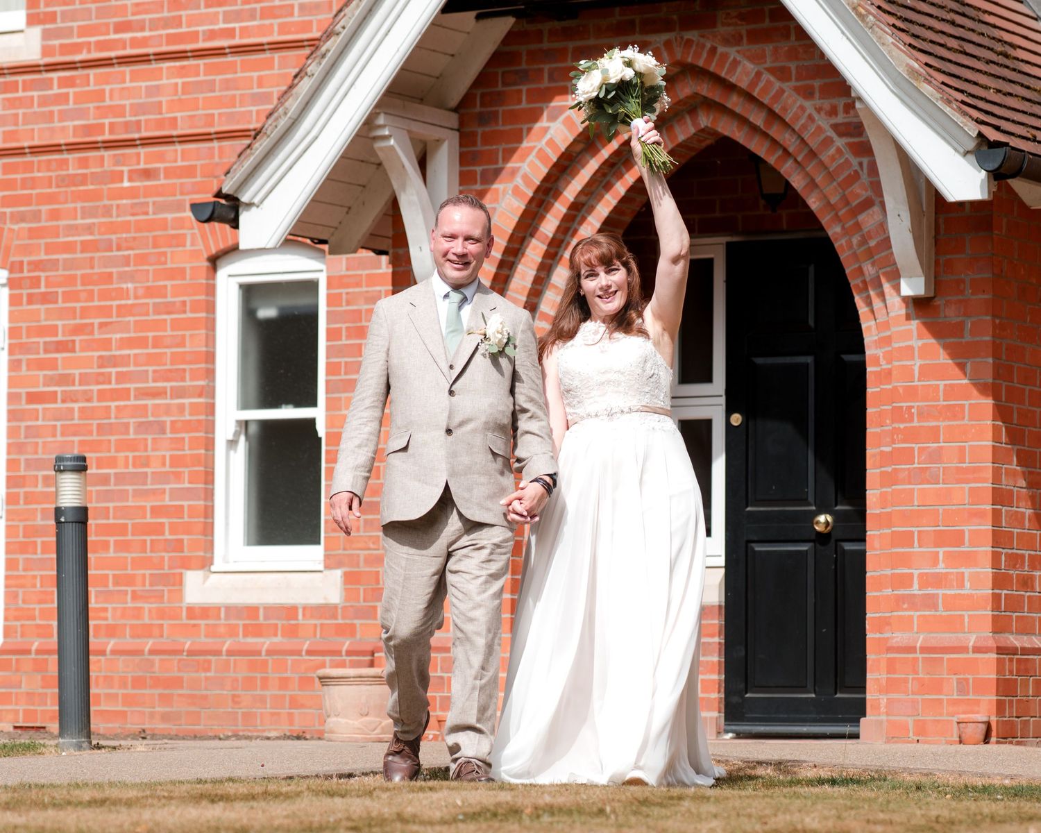 Happy newlyweds celebrate outside a red brick church, bride in white gown holding bouquet aloft, groom in tan suit.