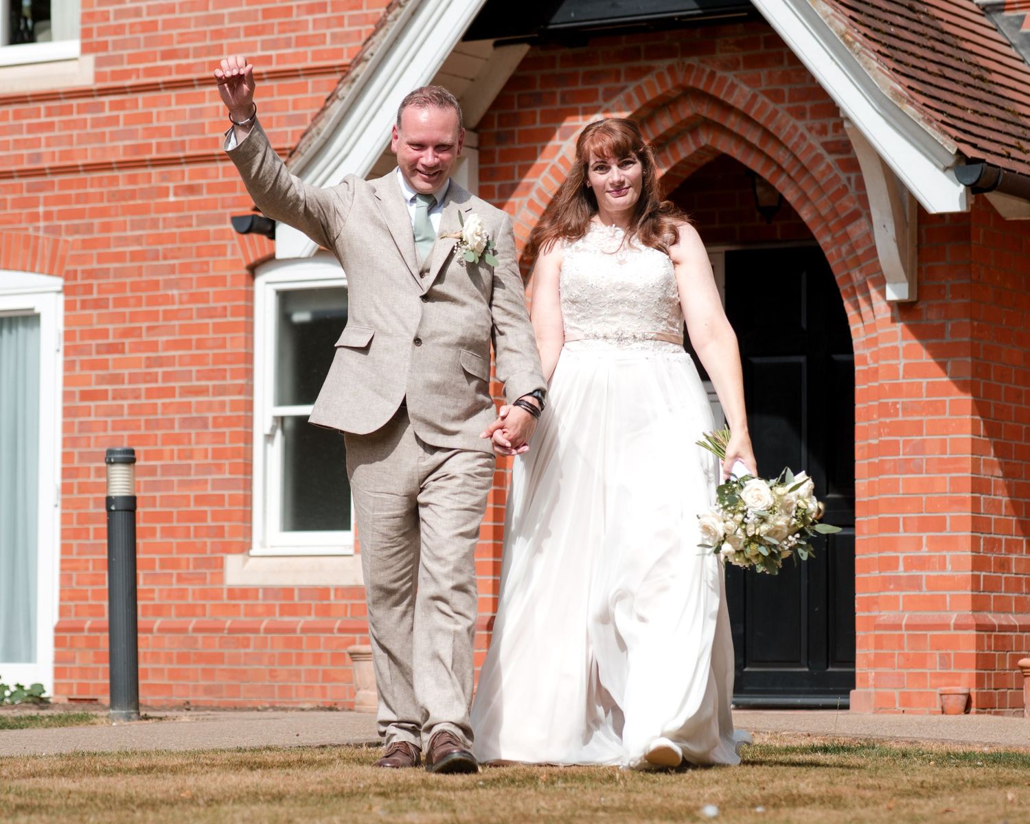 Happy newlyweds holding hands outside a red brick building, groom in tan suit waving, bride in white gown holding bouquet.