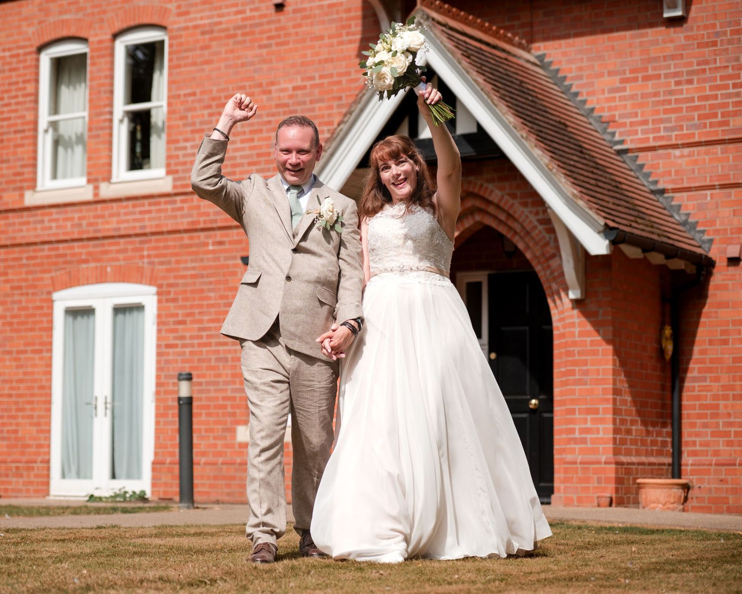Joyful newlywed couple celebrating outside a red brick building, bride in white gown holding bouquet, groom in gray suit.