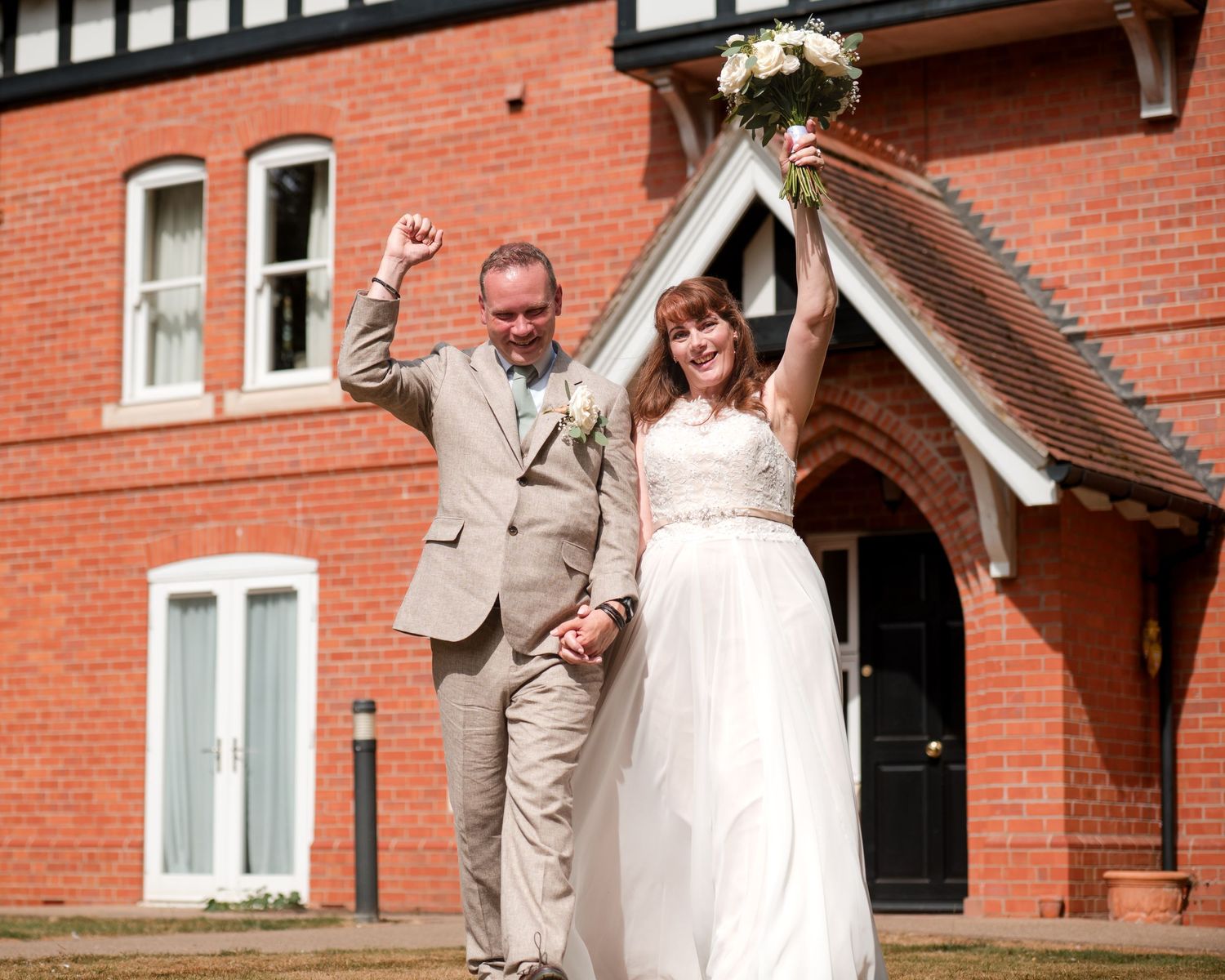Joyful newlyweds celebrate outside a red brick building, bride in white gown holding bouquet, groom in tan suit raising fist.