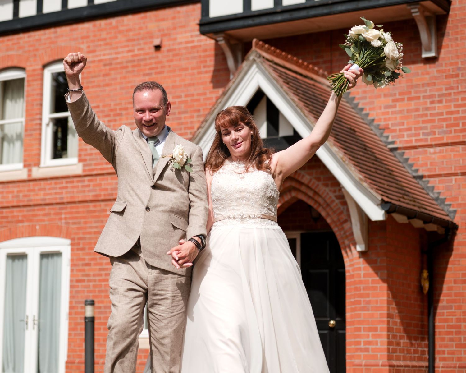 Joyful newlyweds celebrate outside a red brick building, bride holding bouquet and groom raising fist in triumph.