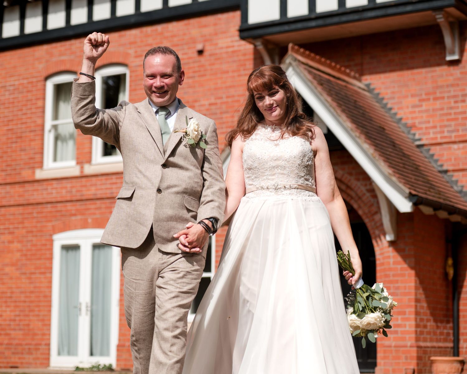 Joyful newlyweds celebrate outside a red brick building, groom raising fist in excitement, bride holding bouquet in white gown.