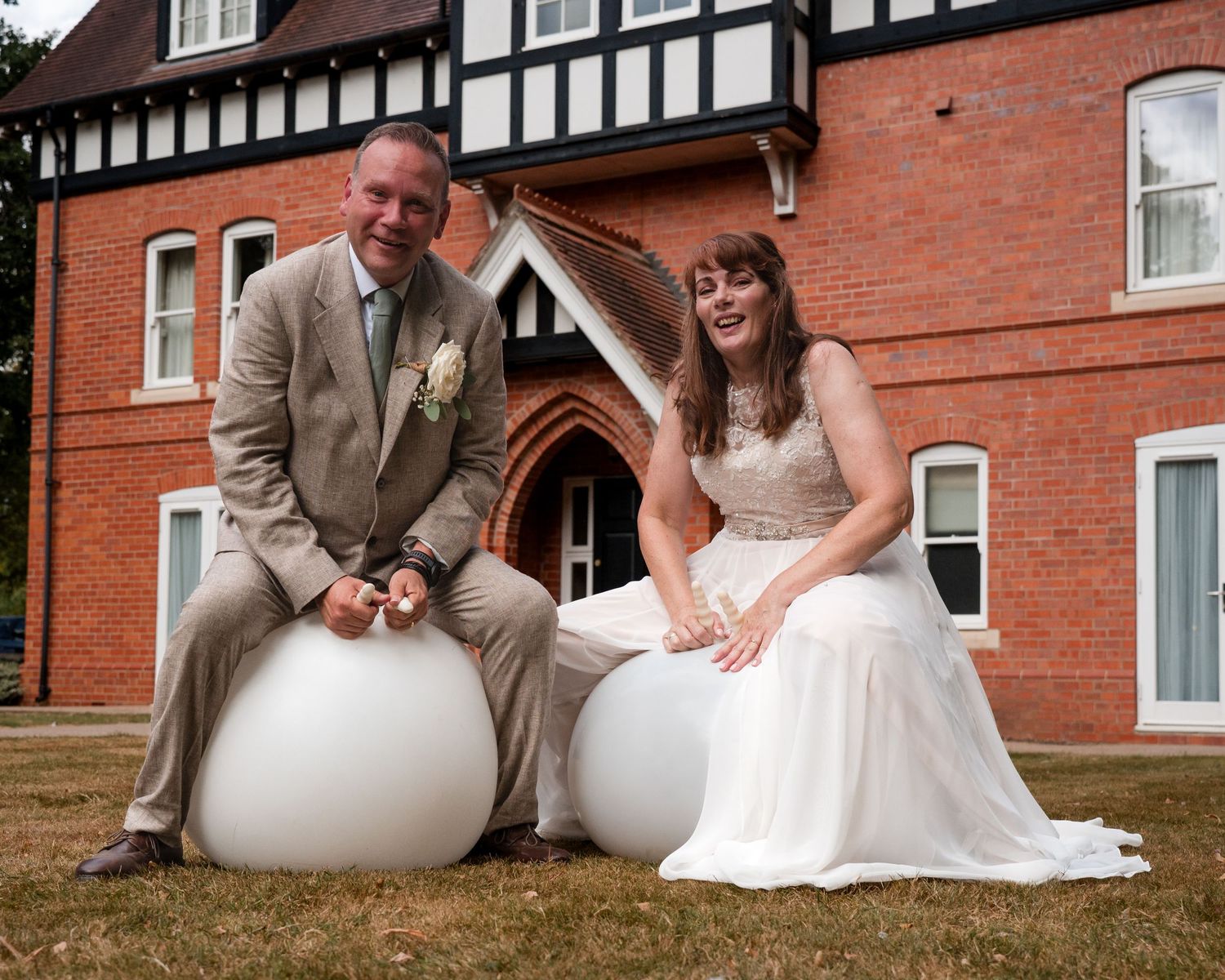 Bride and groom sitting on white round seats outside a red brick Tudor-style venue on their wedding day.