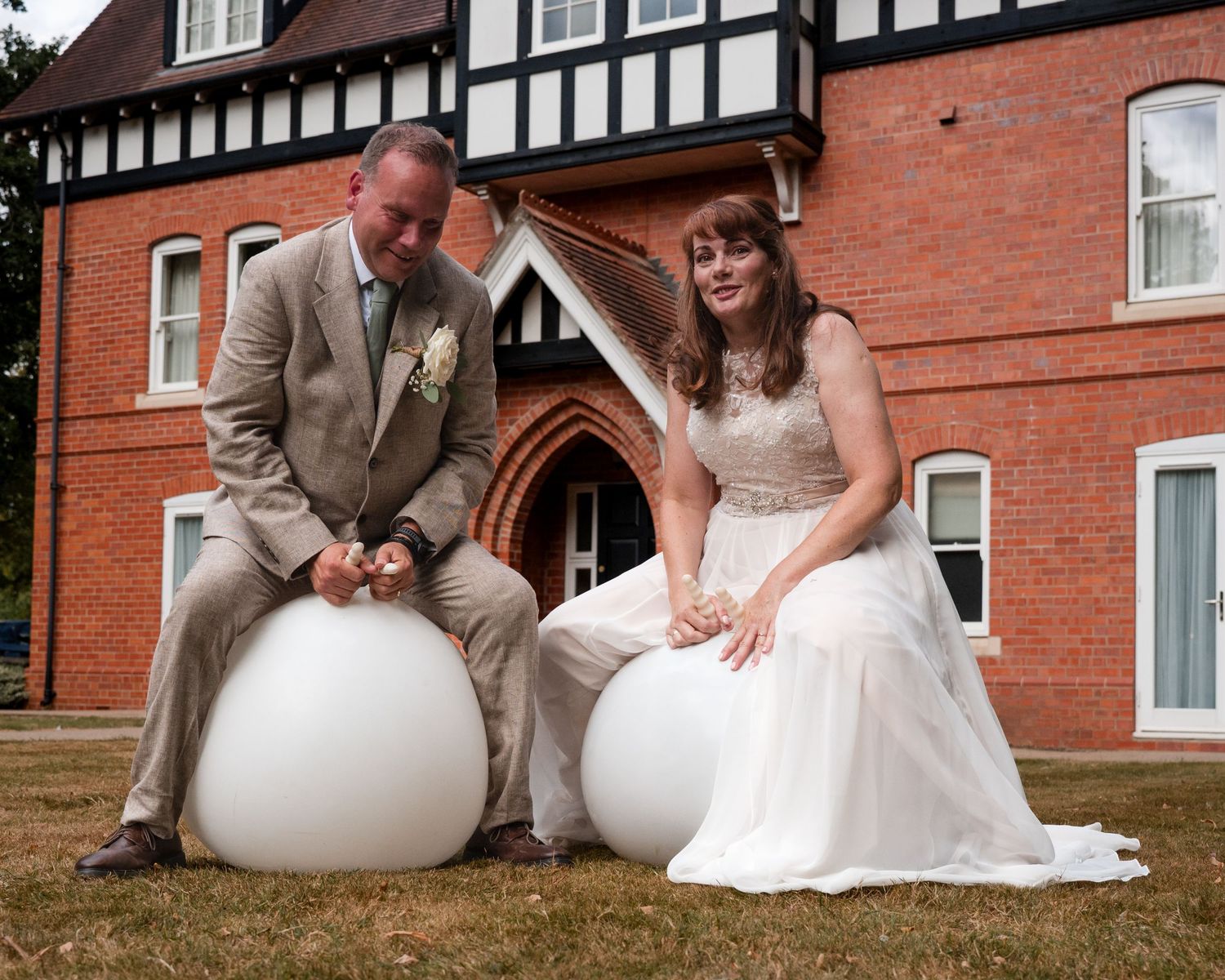 Bride and groom sitting on large white spherical seats outside a Tudor-style brick wedding venue.