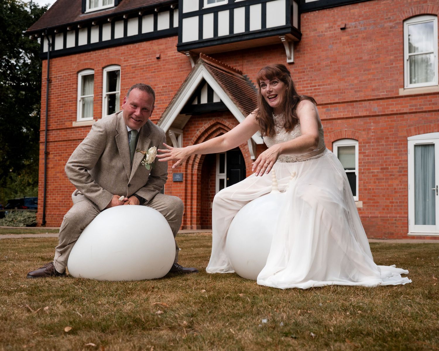 Bride and groom posing with white egg-shaped seats on lawn outside red brick Tudor-style wedding venue.