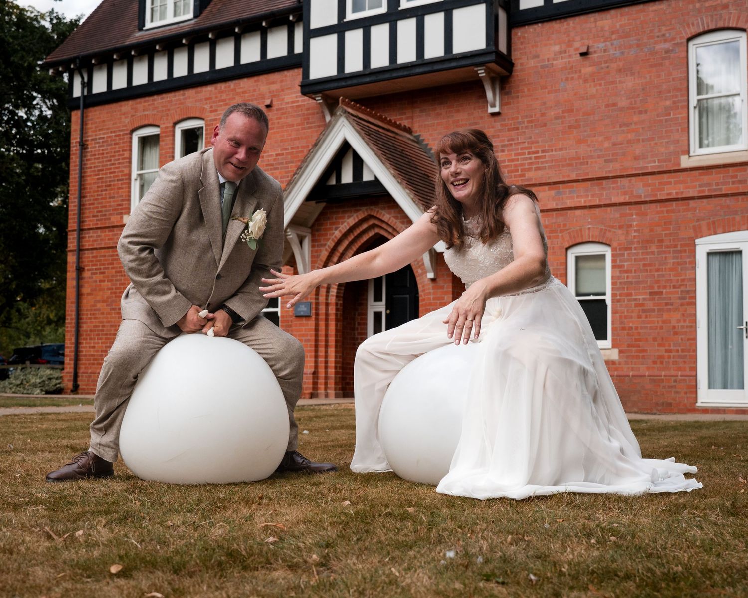 Bride and groom sitting on large white spheres outside a red brick Tudor-style building on their wedding day.