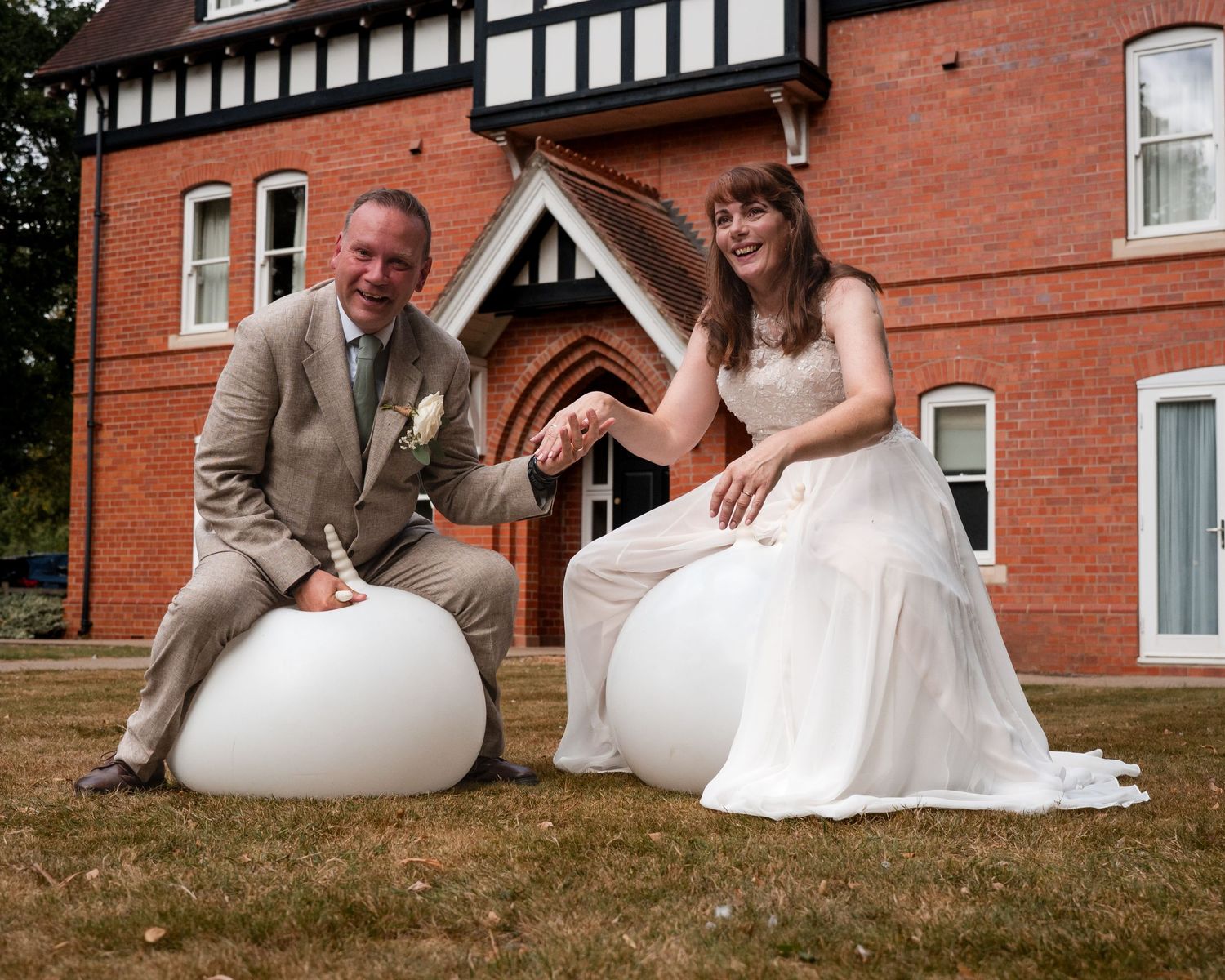 Bride and groom sitting on white round seats outside a red brick building on their wedding day.