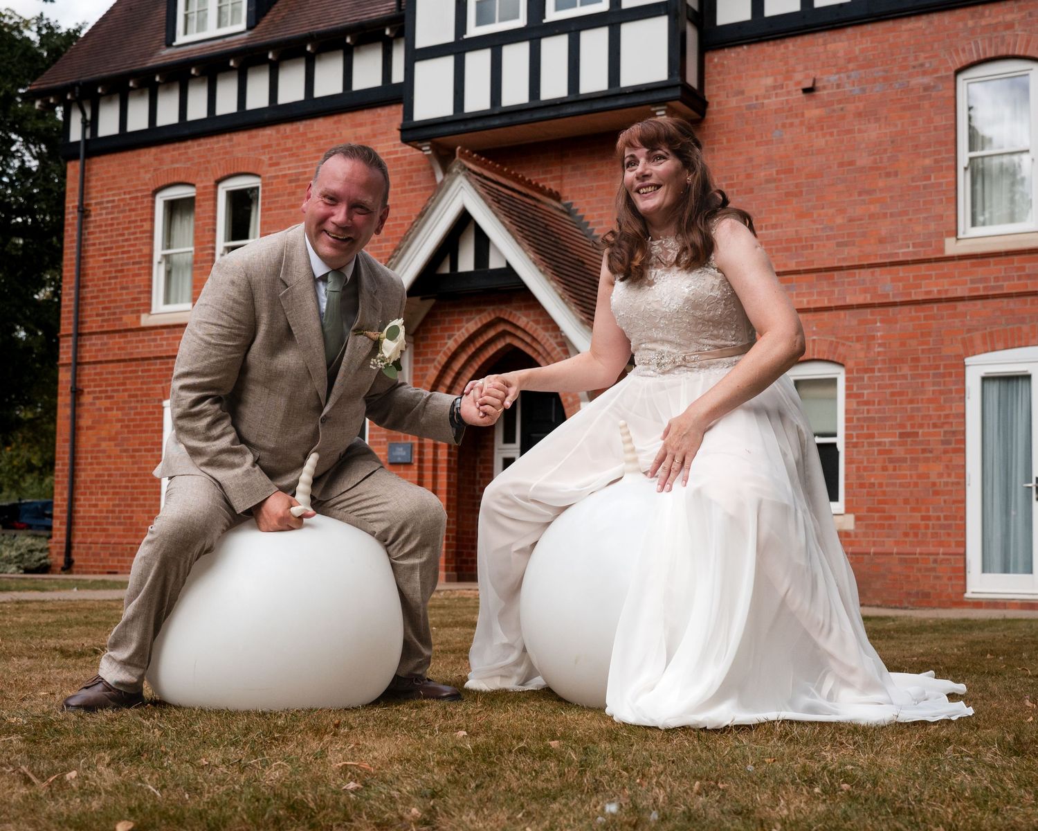Bride and groom sitting on white round seats outside a Tudor-style brick building on their wedding day.
