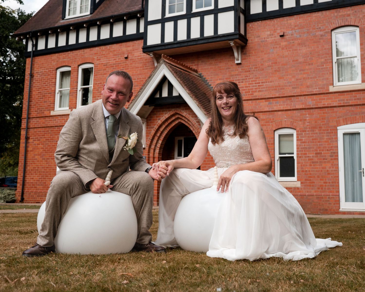Bride and groom sitting on white globe chairs outside a red brick Tudor-style building on their wedding day.