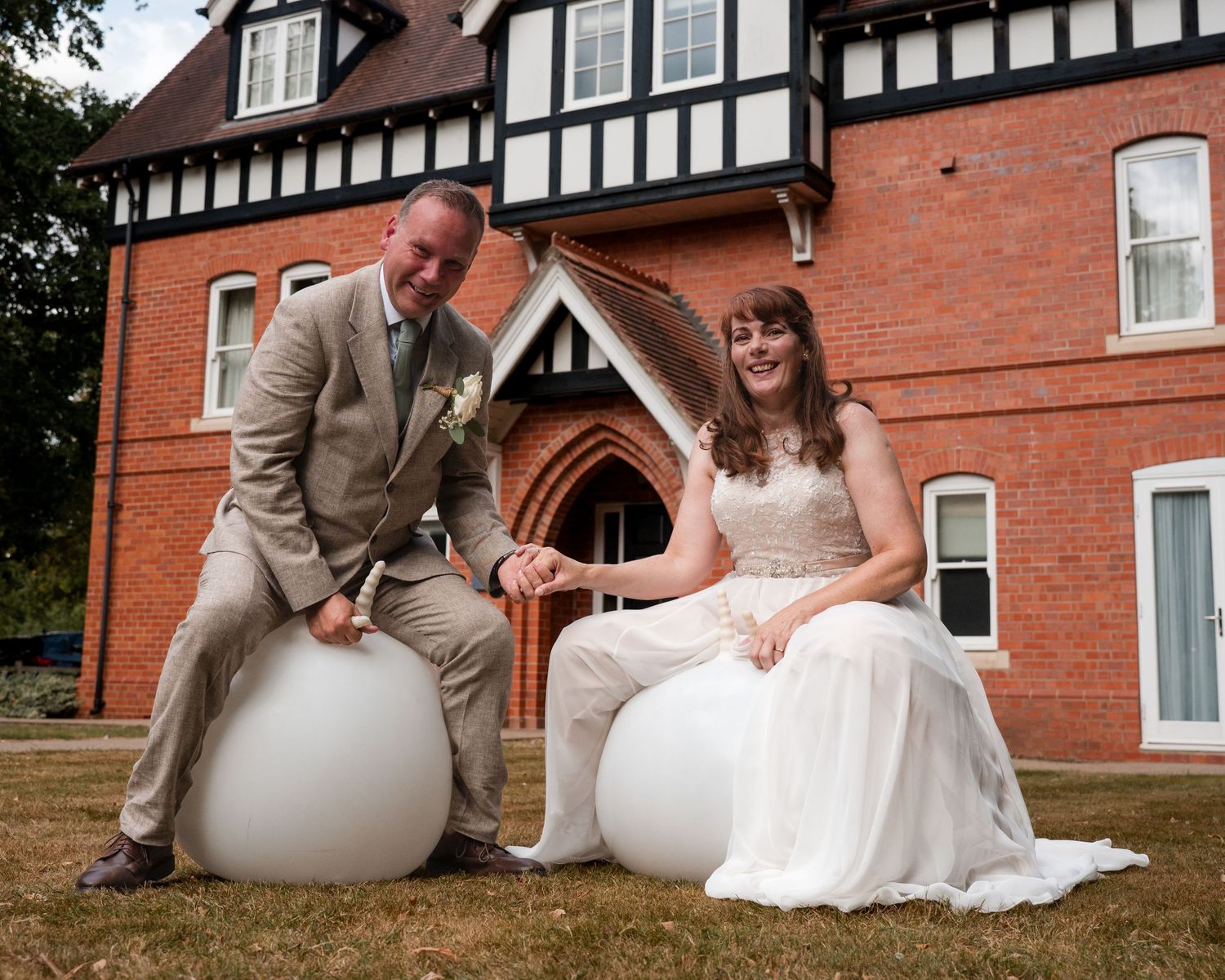 Bride and groom sitting on white spherical seats outside a Tudor-style brick building on their wedding day.