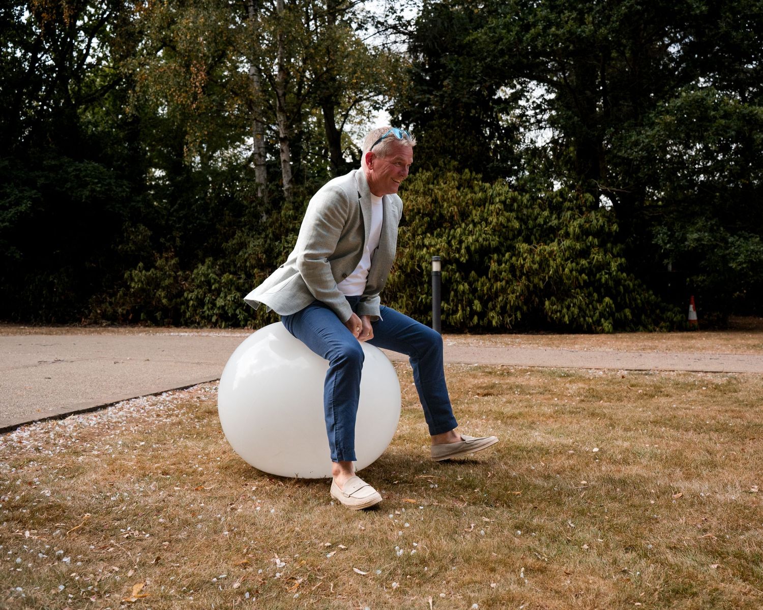 Older man with gray hair sits on a large white decorative ball outdoors on gravel near green trees.