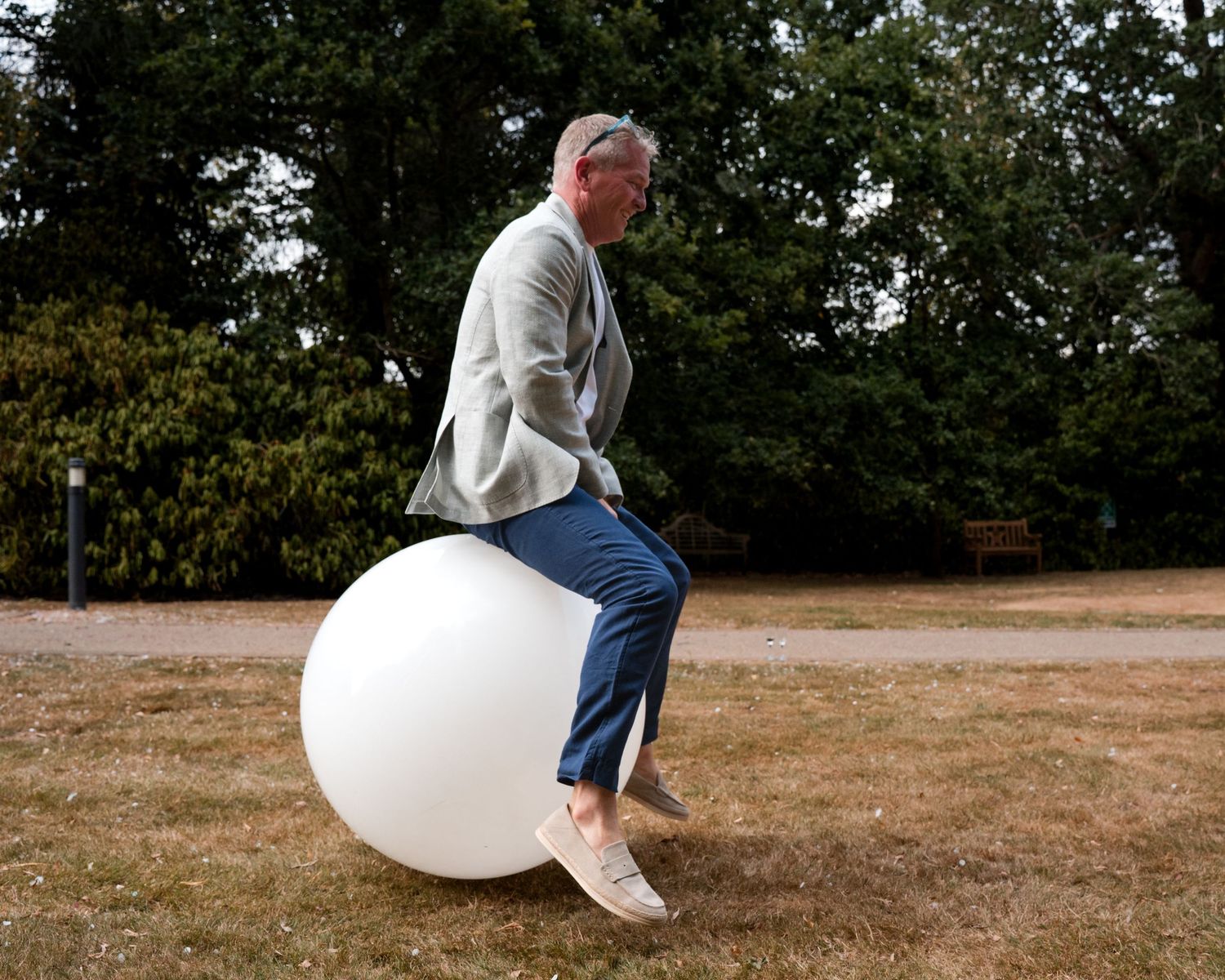 An older man sits on a large white exercise ball outdoors in a park surrounded by green trees.