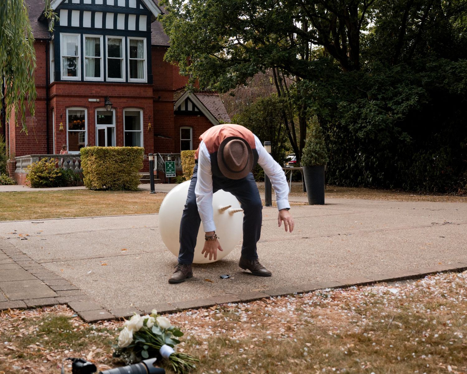 Man in hat bending over large white sphere on paved path outside red brick building with flowers nearby.
