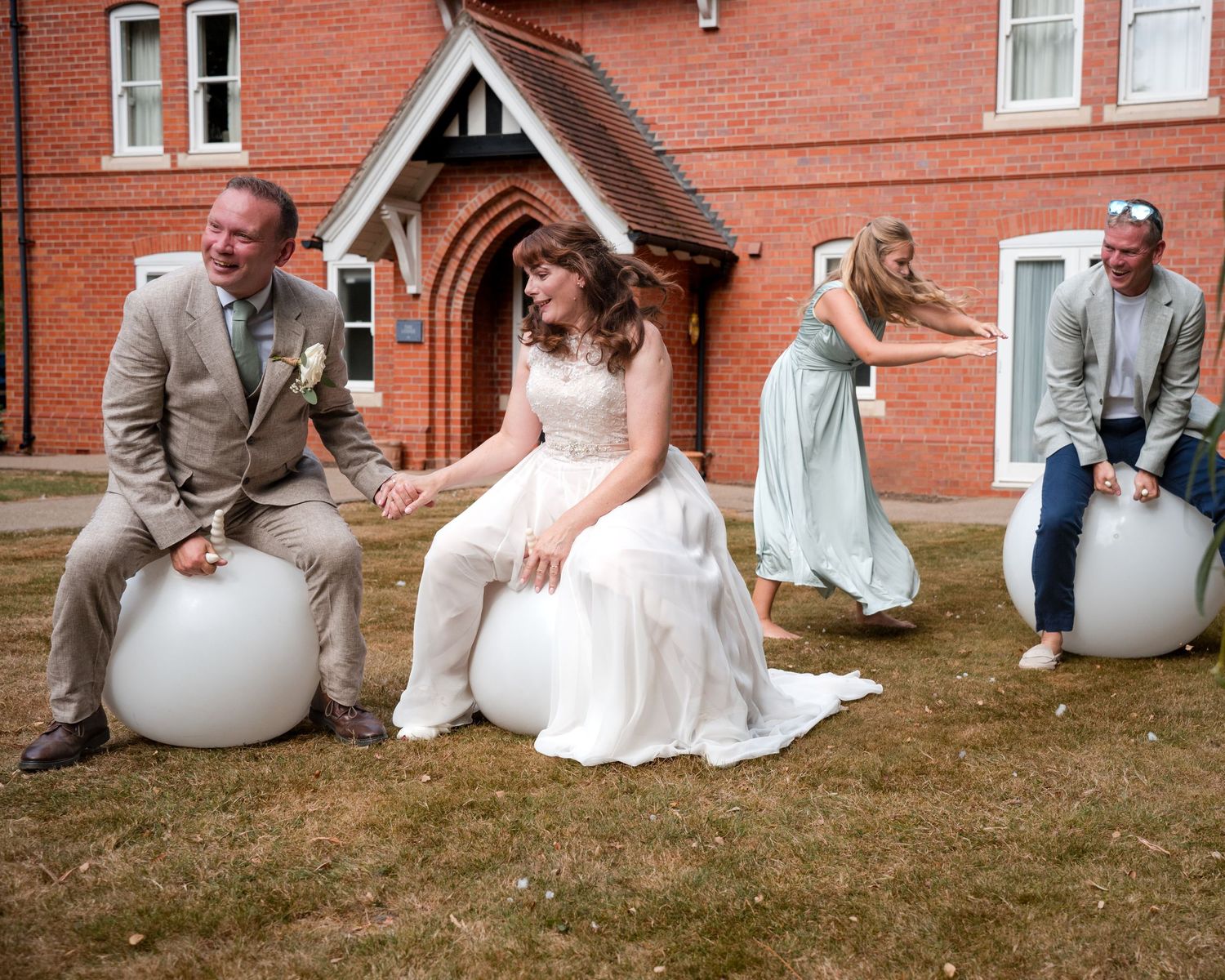 Bride and groom bounce on space hoppers outside a red brick building during a fun outdoor wedding celebration.