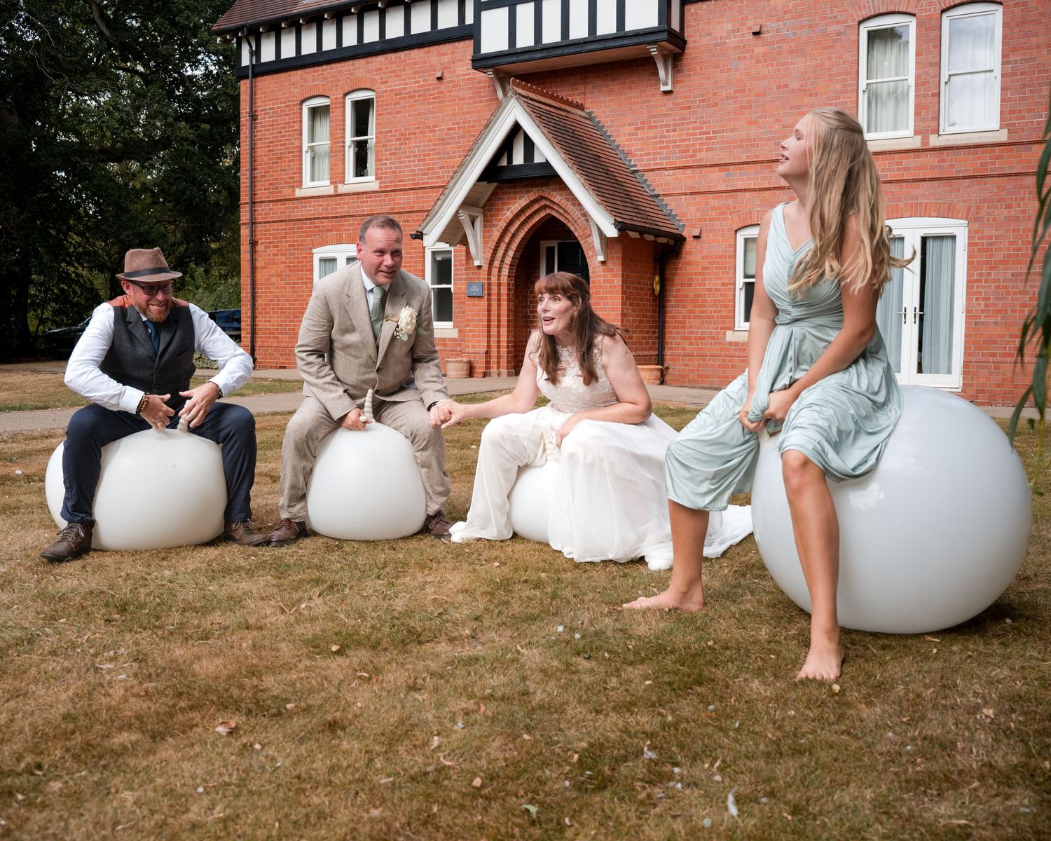Wedding party sitting on large white spherical seats outside a red brick Tudor-style building on a sunny day.