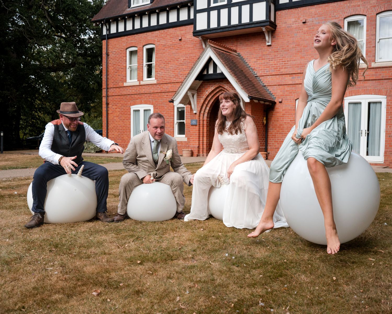 Wedding party laughing on white round outdoor seats in front of a red brick Tudor-style mansion on a sunny day.